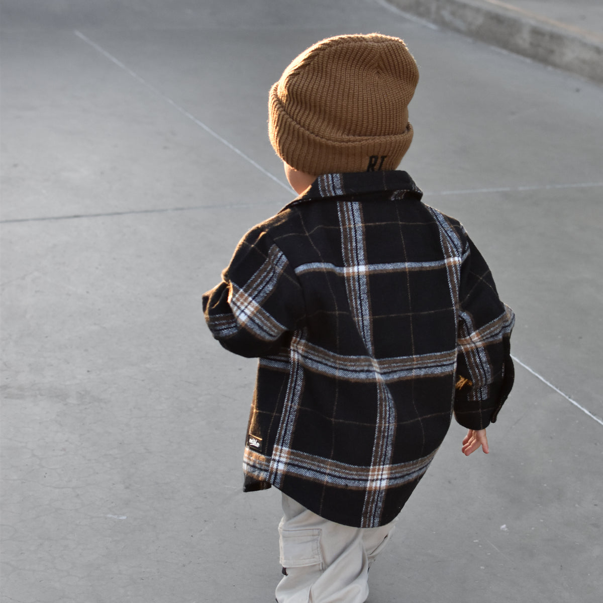 A toddler viewed from behind wearing a tan knit beanie, a black and white plaid flannel shirt, light beige pants, and black and white sneakers, standing on a concrete skatepark surface.