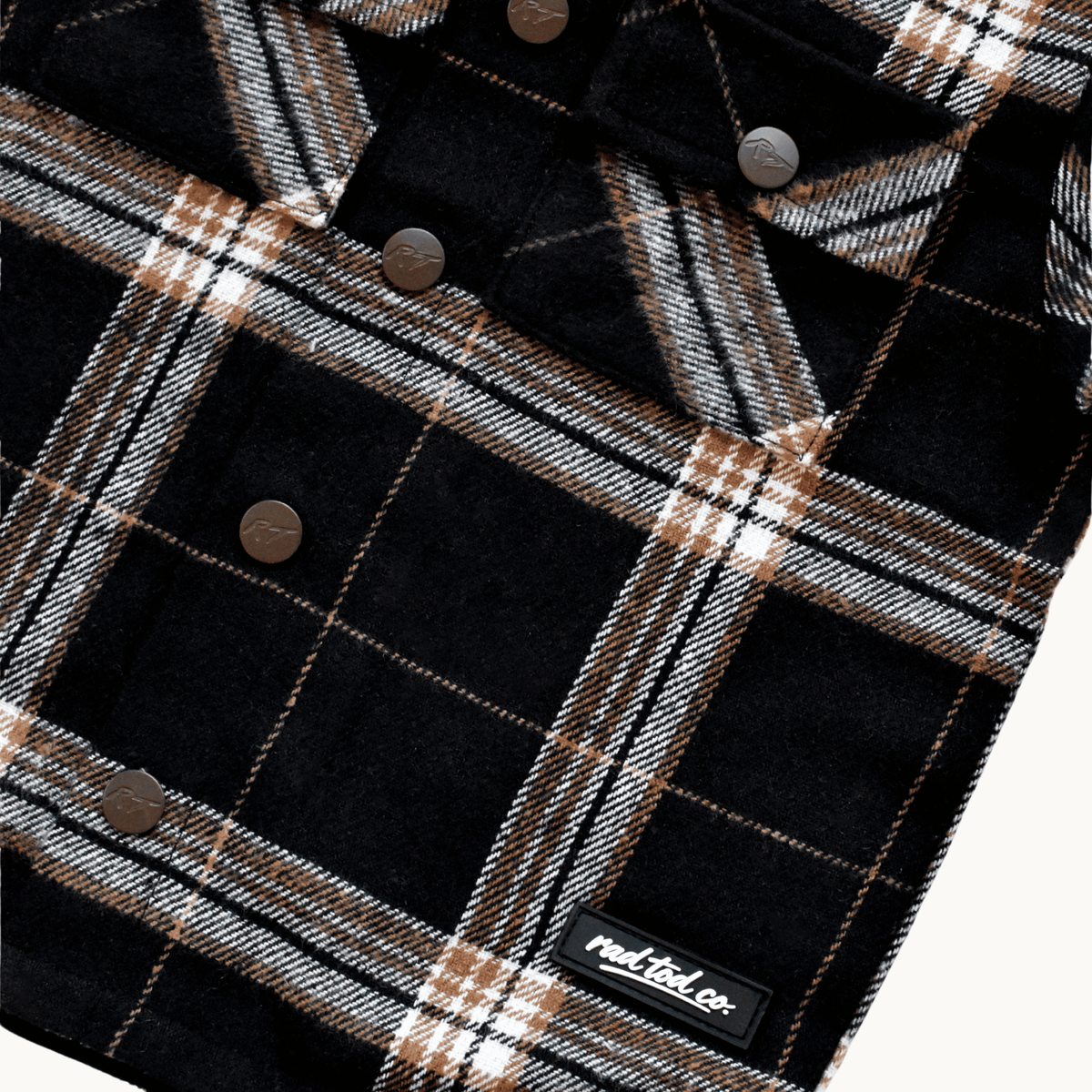 Close-up of a black plaid flannel shirt jacket with brown buttons and a small 'RAD TOD co.' label near the bottom right, shown on a light background.
