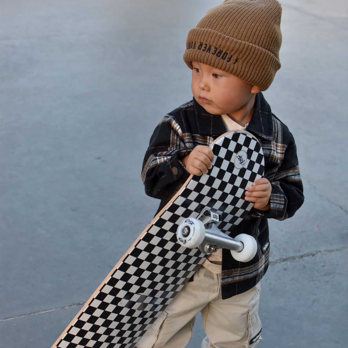 A toddler stands outdoors holding a skateboard with a black-and-white checkerboard deck. The child wears a tan knit beanie with dark lettering, a dark plaid jacket, light beige pants, and black-and-white sneakers.