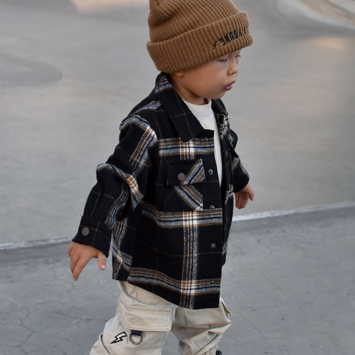A toddler walking in a concrete skatepark wearing a brown knit beanie, a black and beige plaid button-up shirt, beige cargo pants, and black and white sneakers.