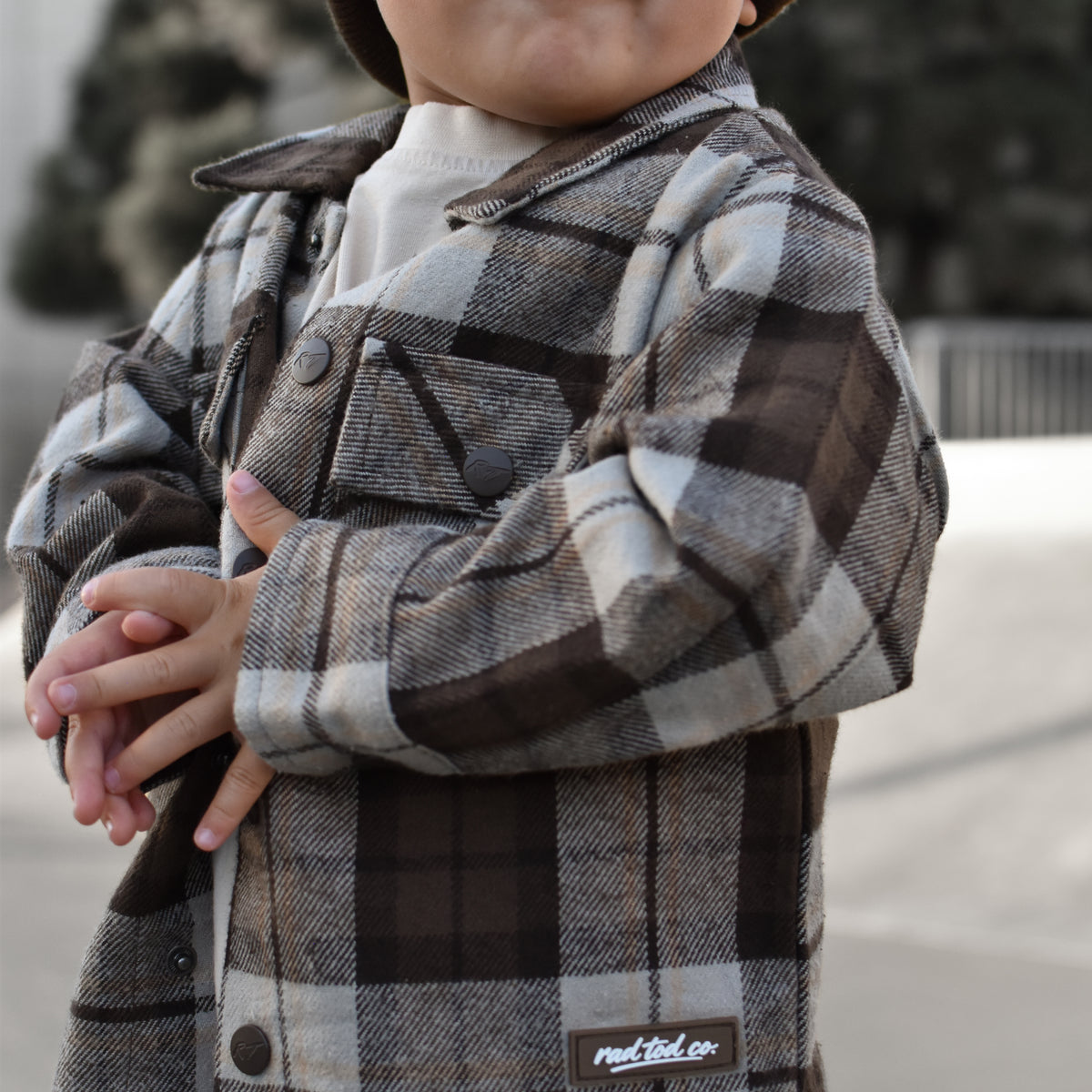 A toddler wearing a brown and beige plaid flannel button-up shirt jacket over a white t-shirt, paired with a matching plaid beanie. The jacket features a visible 'RAD TODDLER logo.
