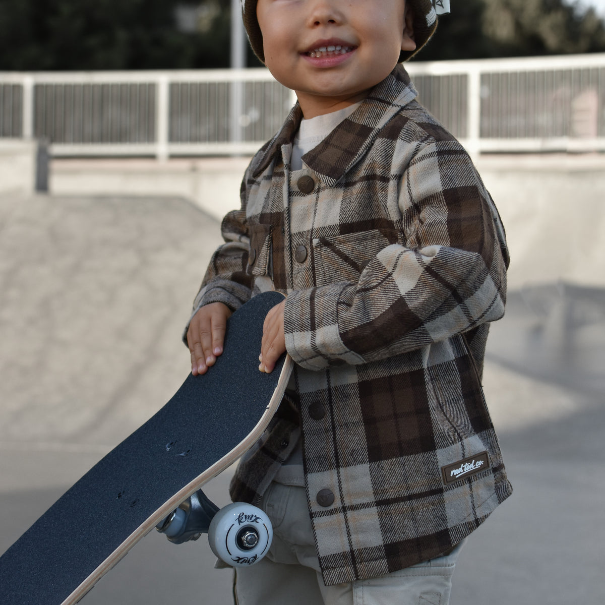 A young child wearing a brown and beige plaid button-up shirt, matching pants, and a checkered beanie hat, holding a skateboard at an outdoor skatepark.