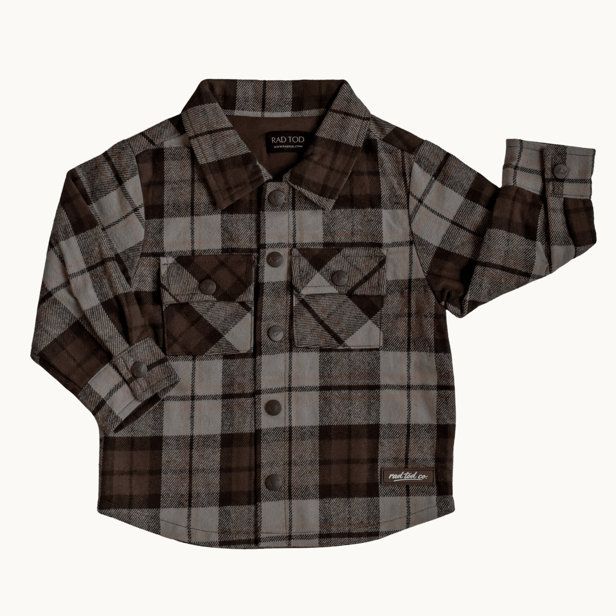 A long-sleeve plaid button-up shirt in brown, beige, and gray, featuring two chest pockets and a small white 'RAD TODDLER CO' logo on the lower right hem, displayed flat against a light off-white background.