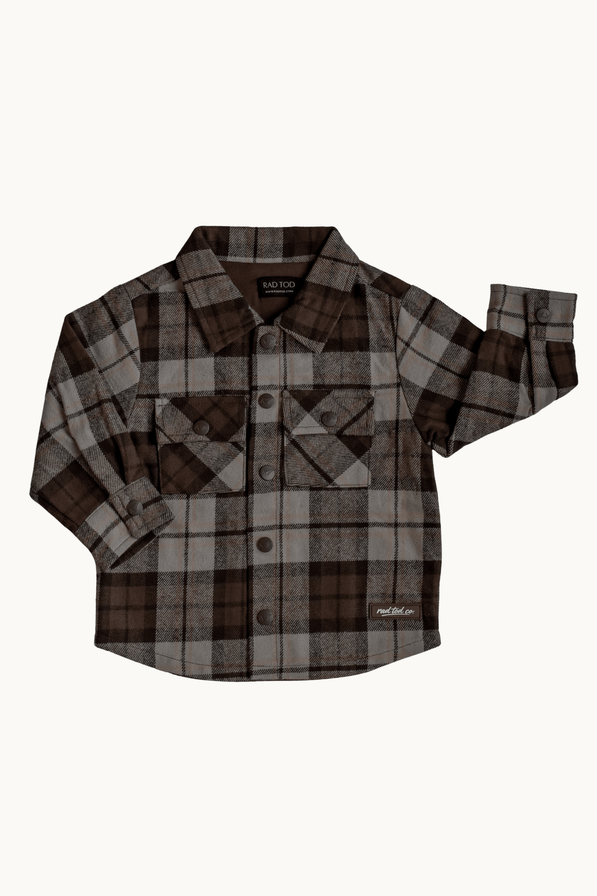 A long-sleeve plaid button-up shirt in brown, beige, and gray, featuring two chest pockets and a small white 'RAD TODDLER CO' logo on the lower right hem, displayed flat against a light off-white background.