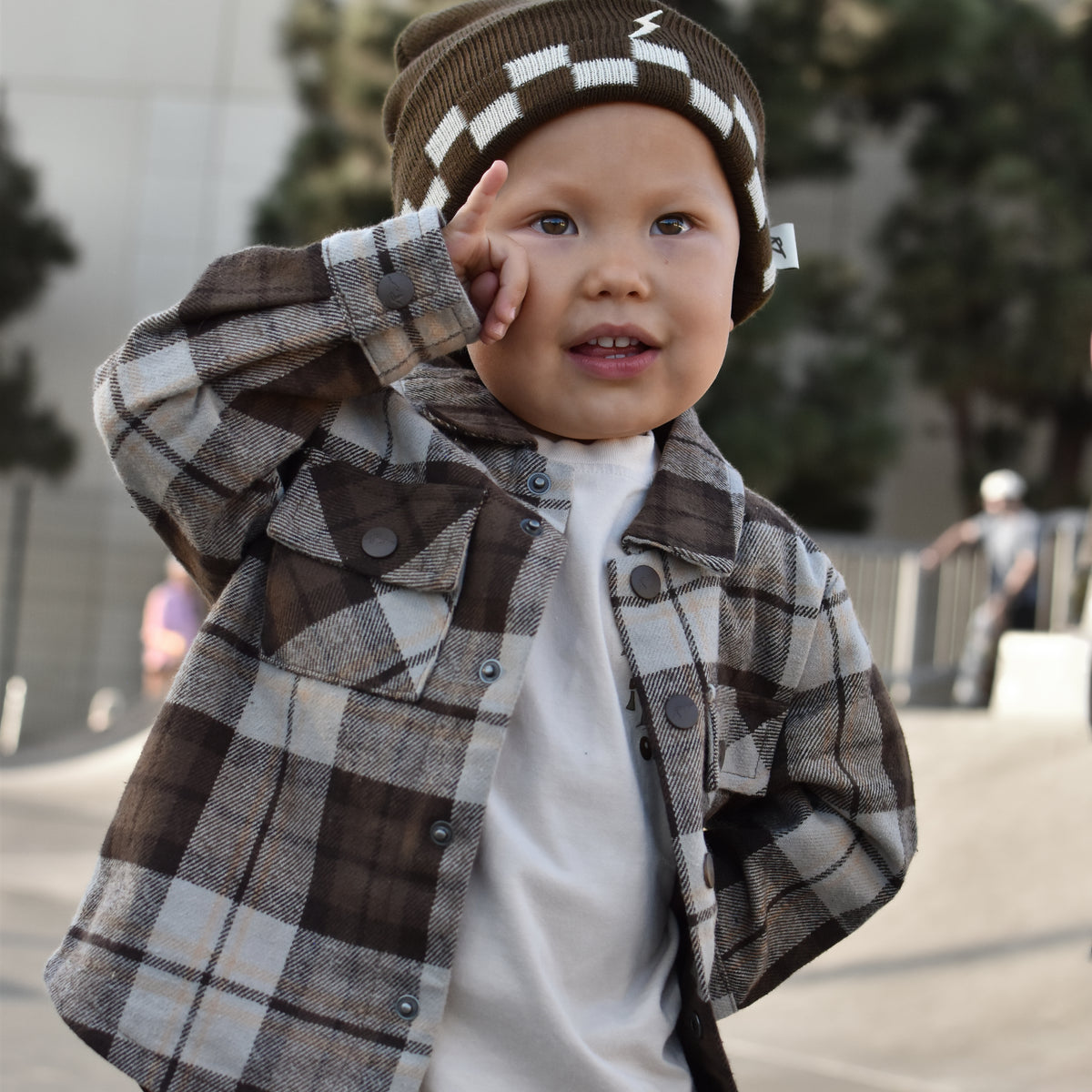 A toddler wearing a brown and beige plaid flannel shirt over a white t-shirt, paired with a brown and white checkered beanie, posing outdoors.
