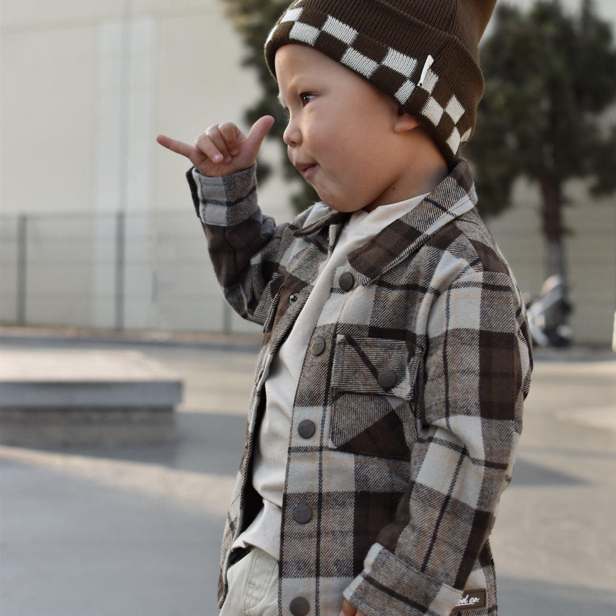 A toddler standing outdoors wearing a brown and beige plaid button-up jacket over a white shirt, paired with a brown knit beanie featuring a white checkerboard pattern.