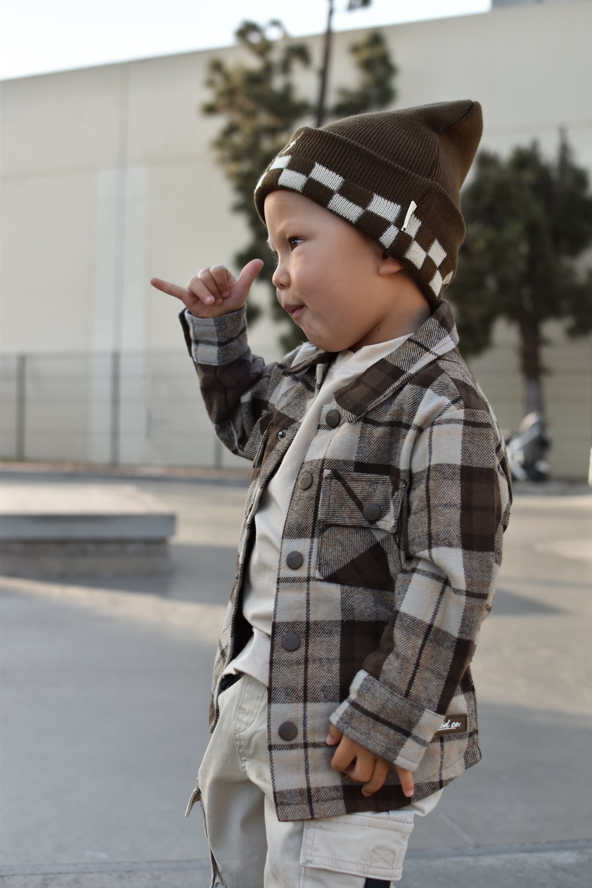 A toddler standing outdoors wearing a brown and beige plaid button-up jacket over a white shirt, paired with a brown knit beanie featuring a white checkerboard pattern.