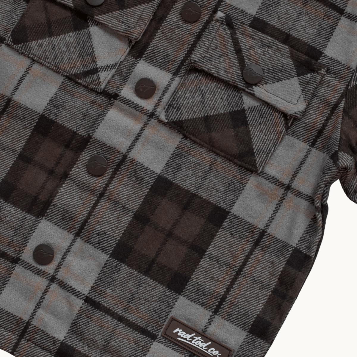A close-up of a plaid flannel shirt jacket in brown, gray, and black, featuring snap-button chest pockets and a visible 'RAD TOD co.' label near the bottom.