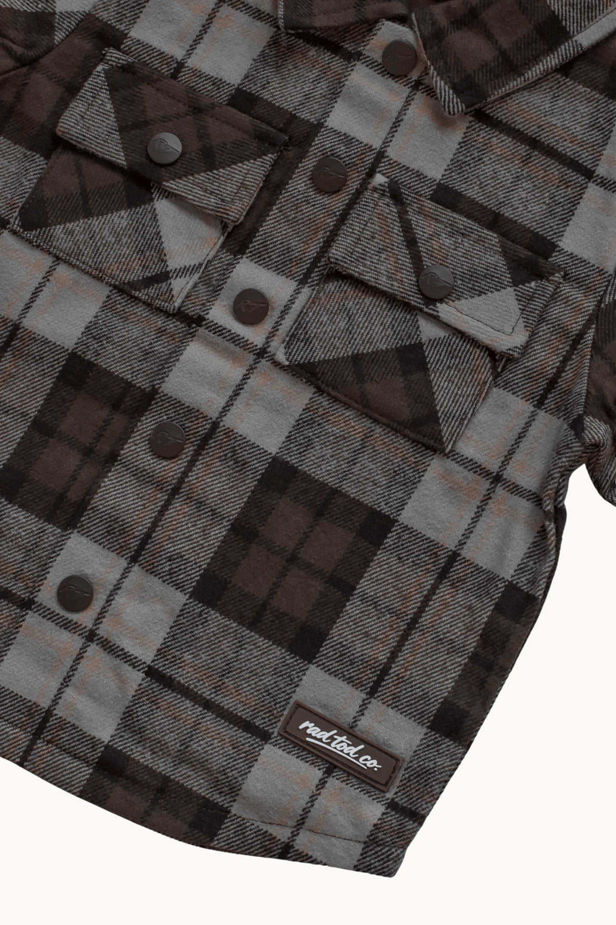 A close-up of a plaid flannel shirt jacket in brown, gray, and black, featuring snap-button chest pockets and a visible 'RAD TOD co.' label near the bottom.