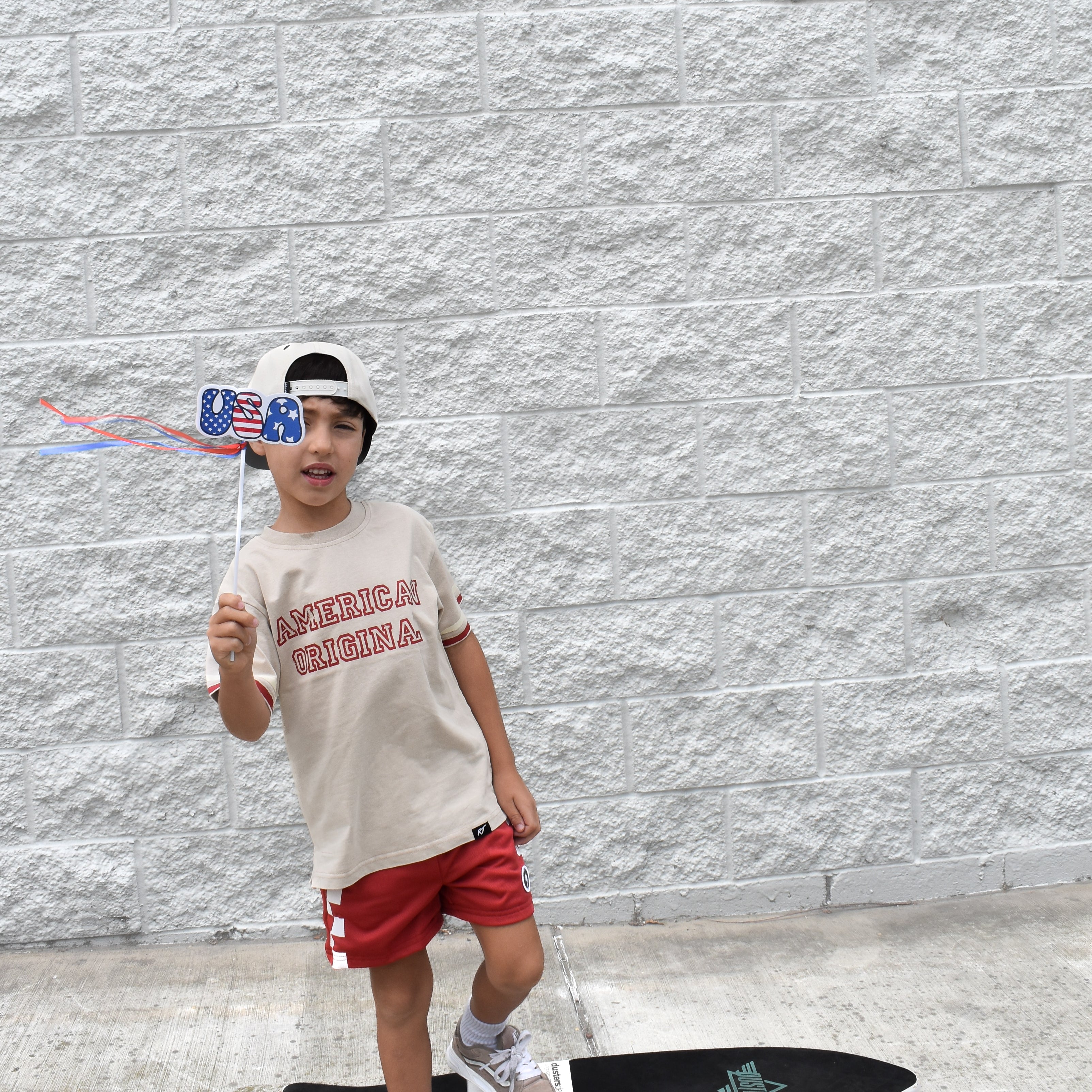 Child with a skateboard and American flag hat standing against a gray wall. Shirt by Rad Toddler