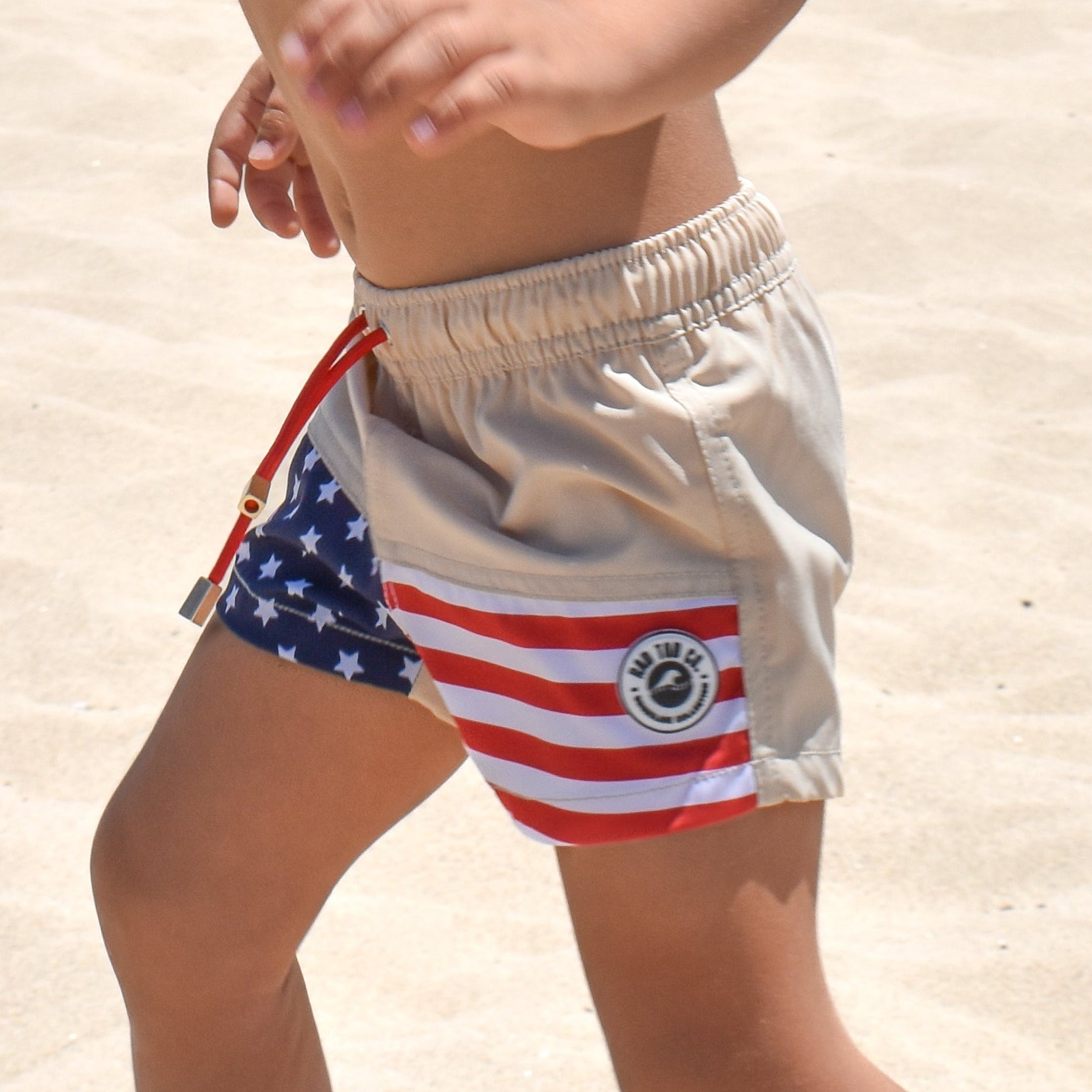Americana toddler swim shorts featuring a beige upper, red and white stripes, blue star pattern, elastic waistband, and vintage-inspired patriotic design for boys.