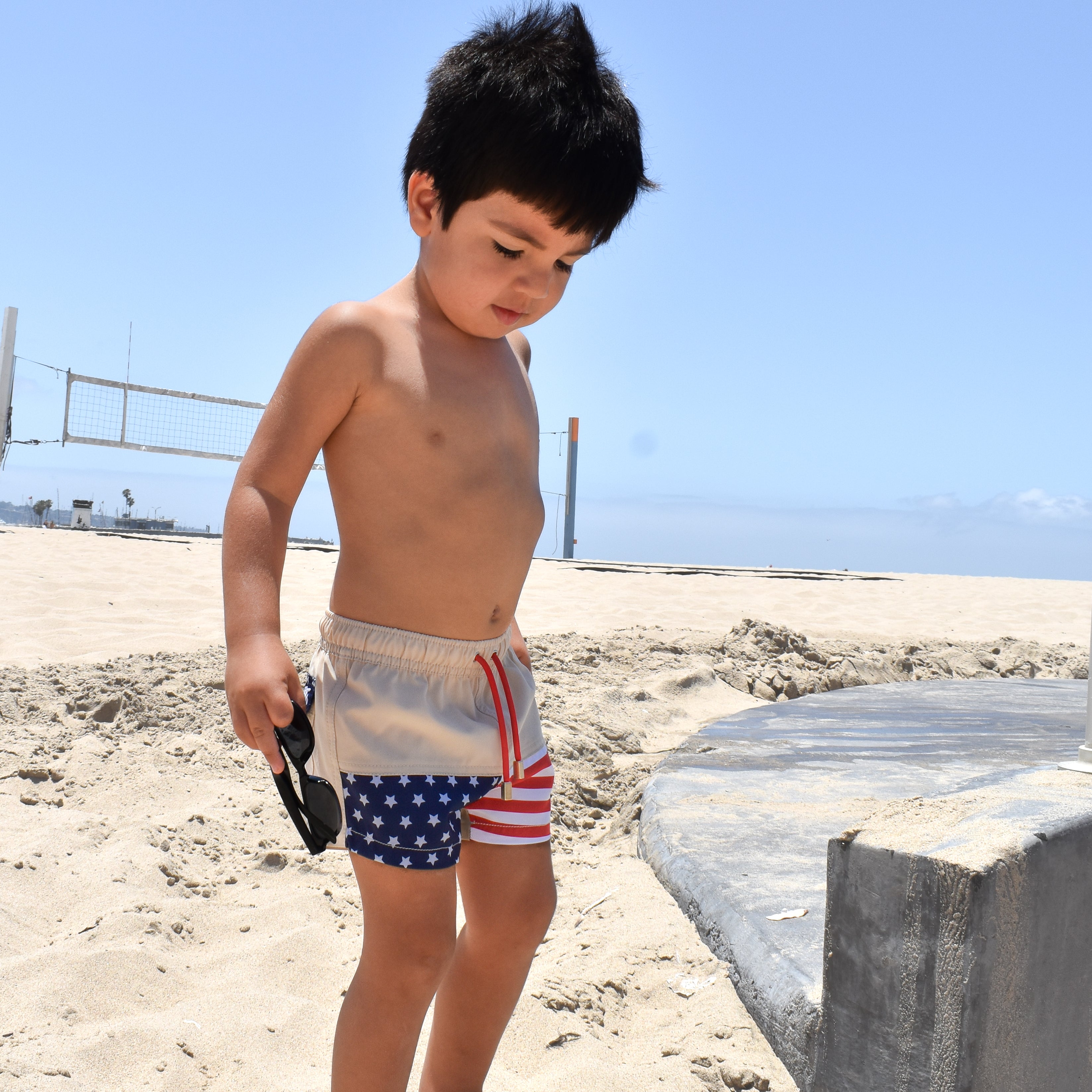 Child in American flag swim shorts standing on a sandy beach with a clear blue sky.