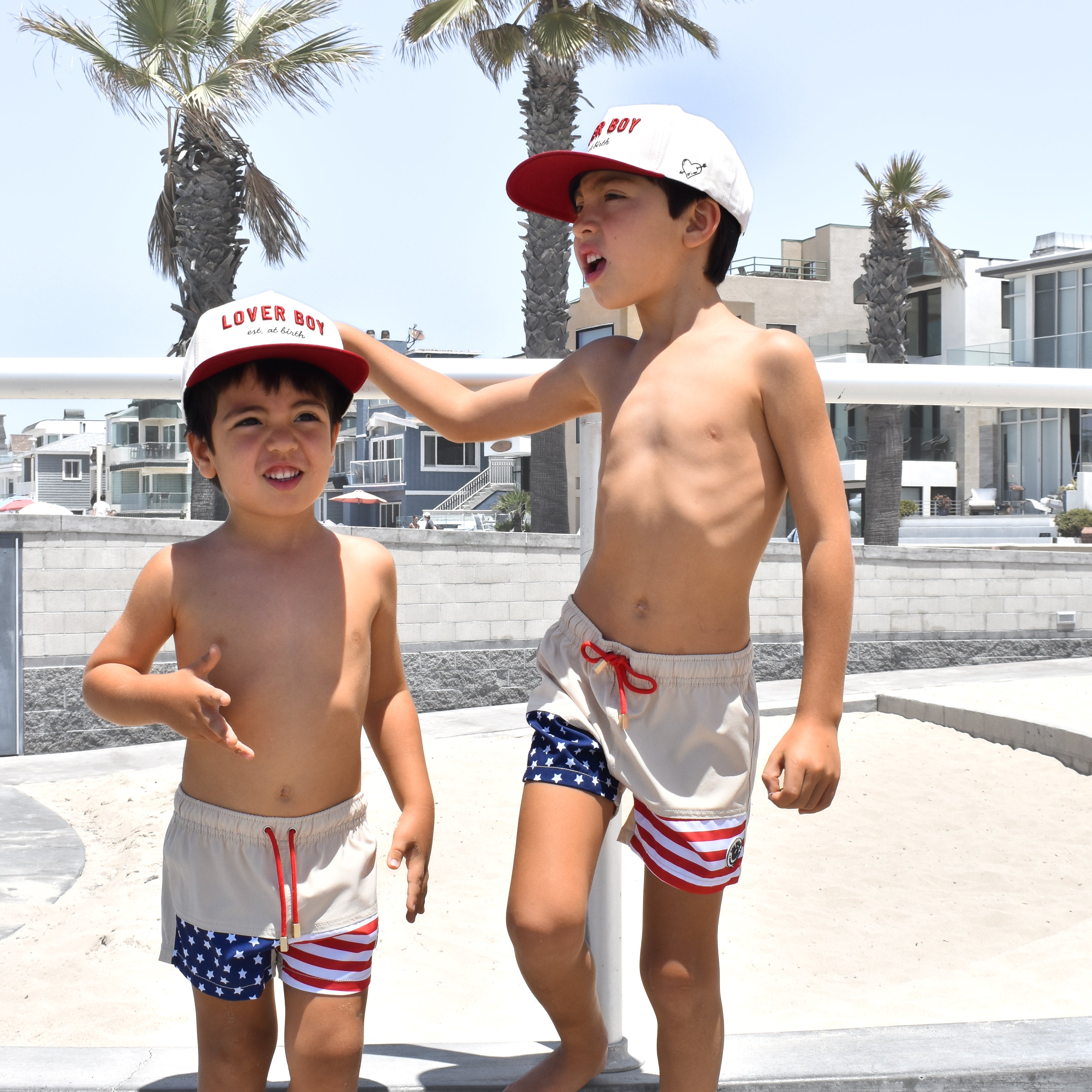 Two children in matching Americana swimsuits and Loverboy hats by RAD TODDLER standing on a concrete surface with palm trees and buildings in the background.
