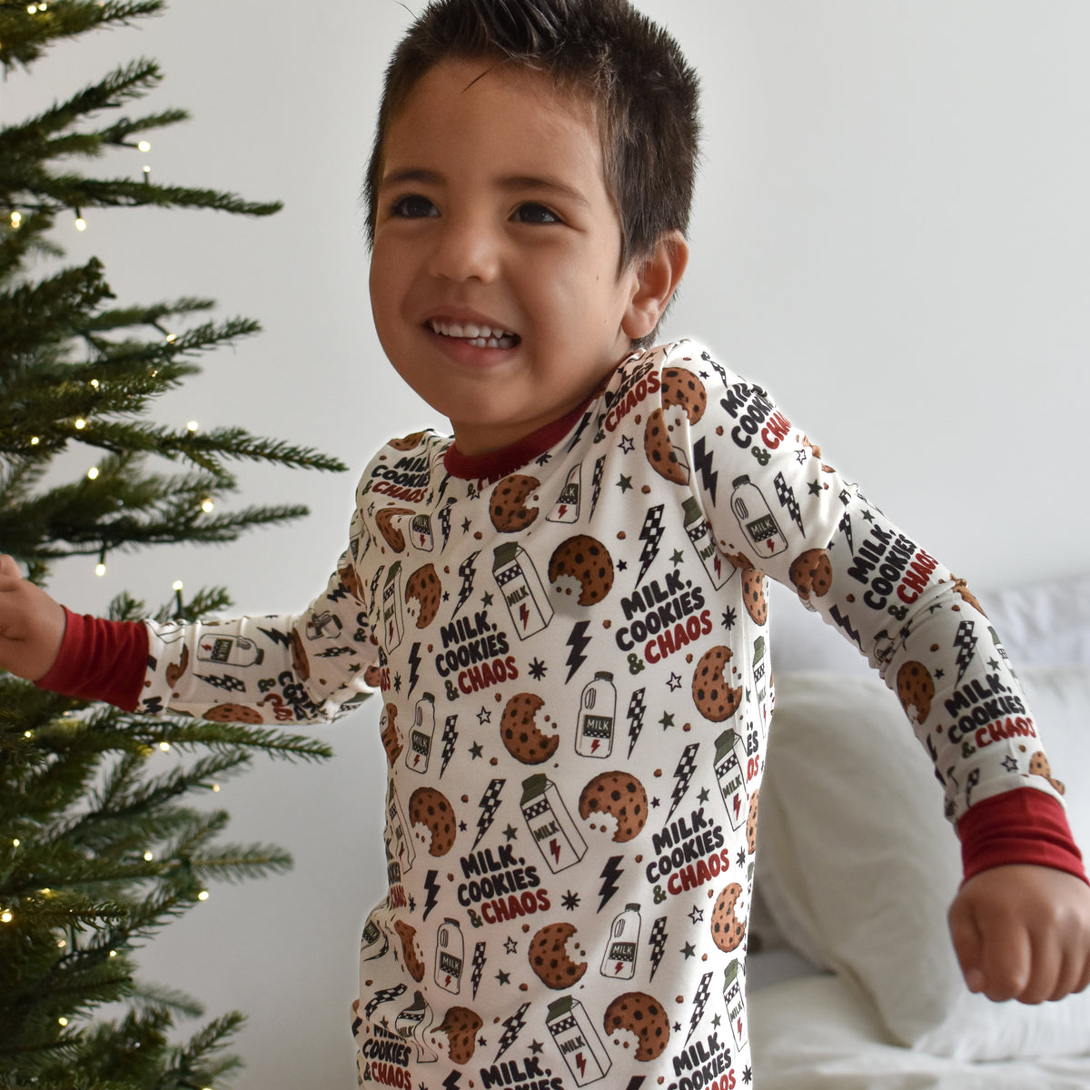 A smiling young boy wearing a long-sleeve Christmas cookie print pajama top and matching pants stands next to a lit Christmas tree in a bedroom setting.
