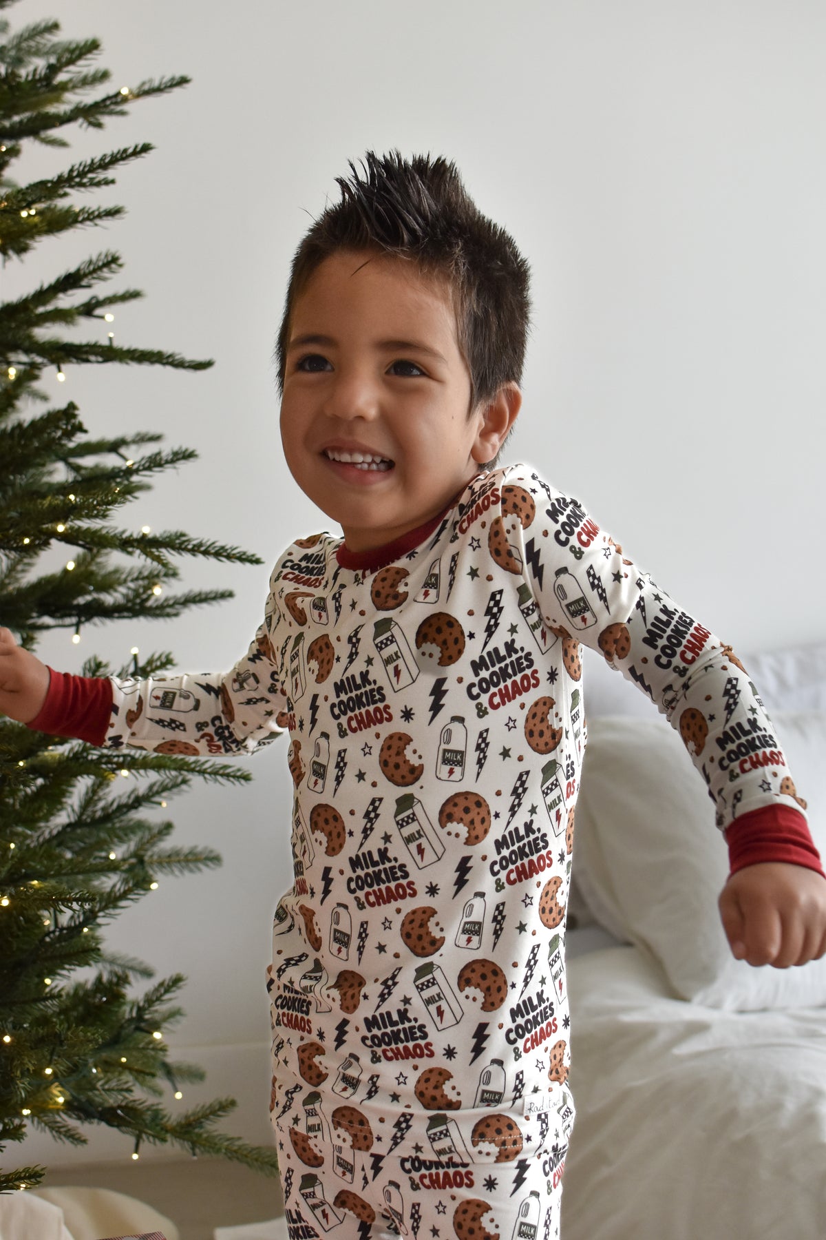 A smiling young boy wearing a long-sleeve Christmas cookie print pajama top and matching pants stands next to a lit Christmas tree in a bedroom setting.