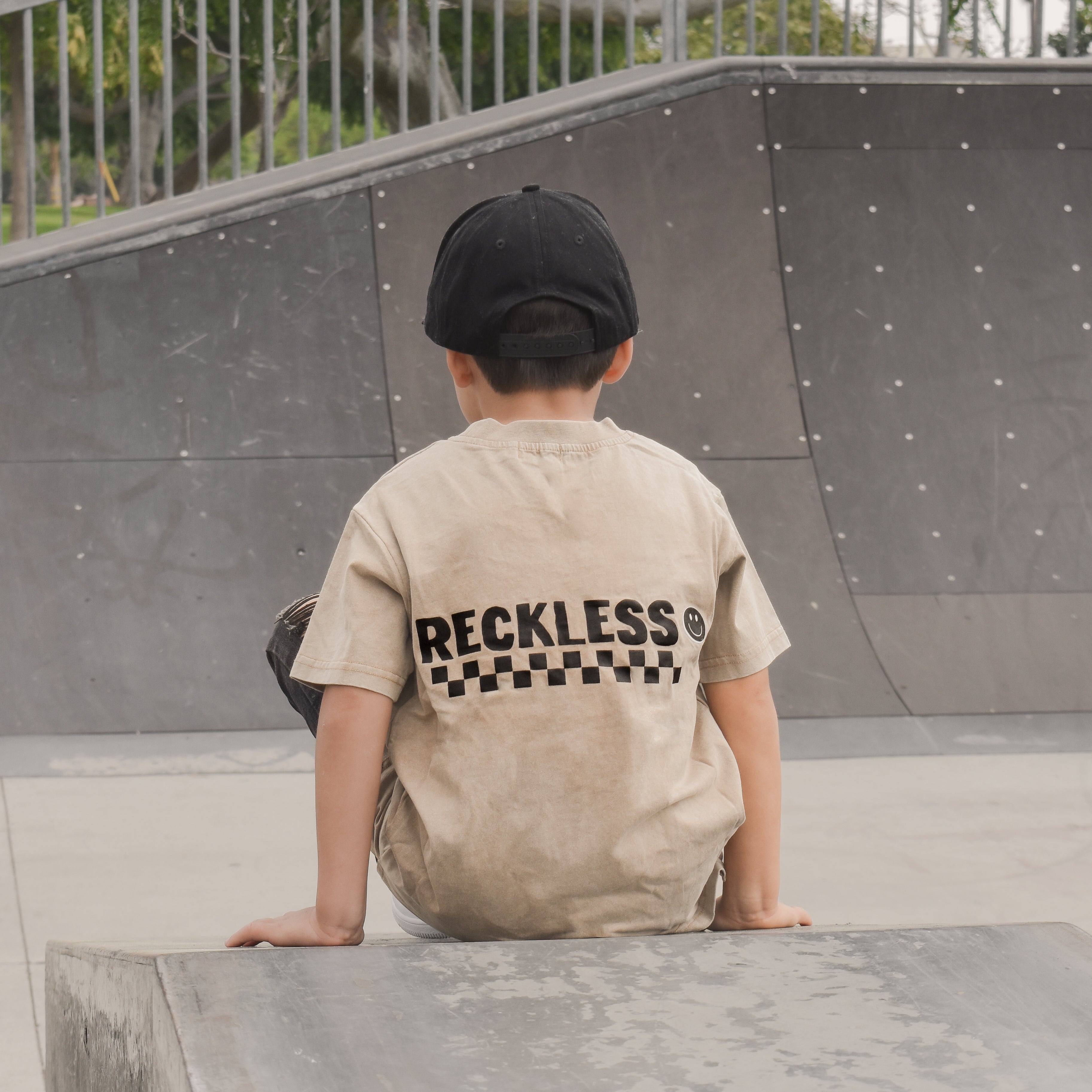 boy wearing Beige acid wash toddler tee with bold 'Reckless' text, checkered patterns, and a smiley face graphic while at the skate park