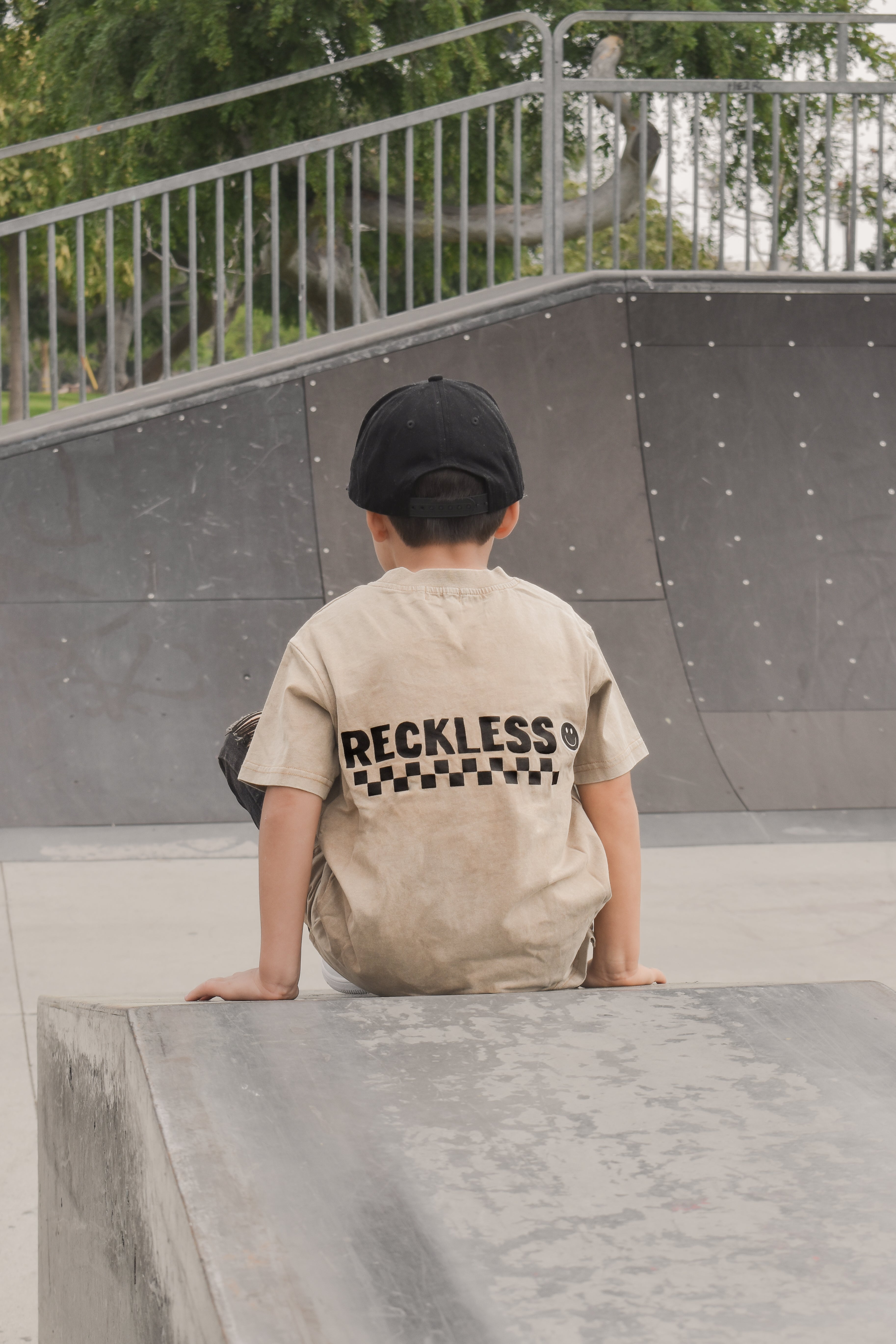 boy wearing Beige acid wash toddler tee with bold 'Reckless' text, checkered patterns, and a smiley face graphic while at the skate park