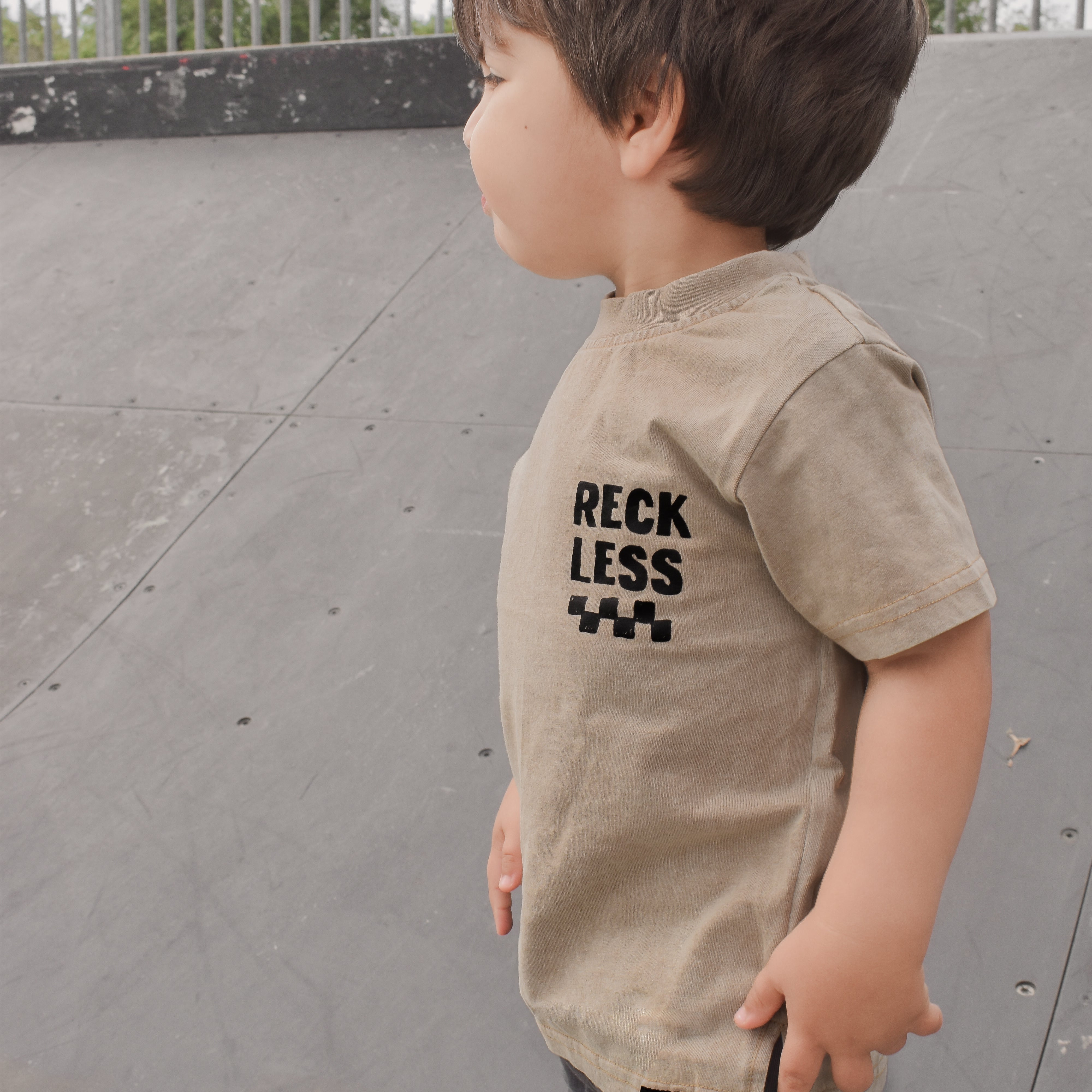 boy wearing Beige acid wash toddler tee with bold 'Reckless' text, checkered patterns, and a smiley face graphic.