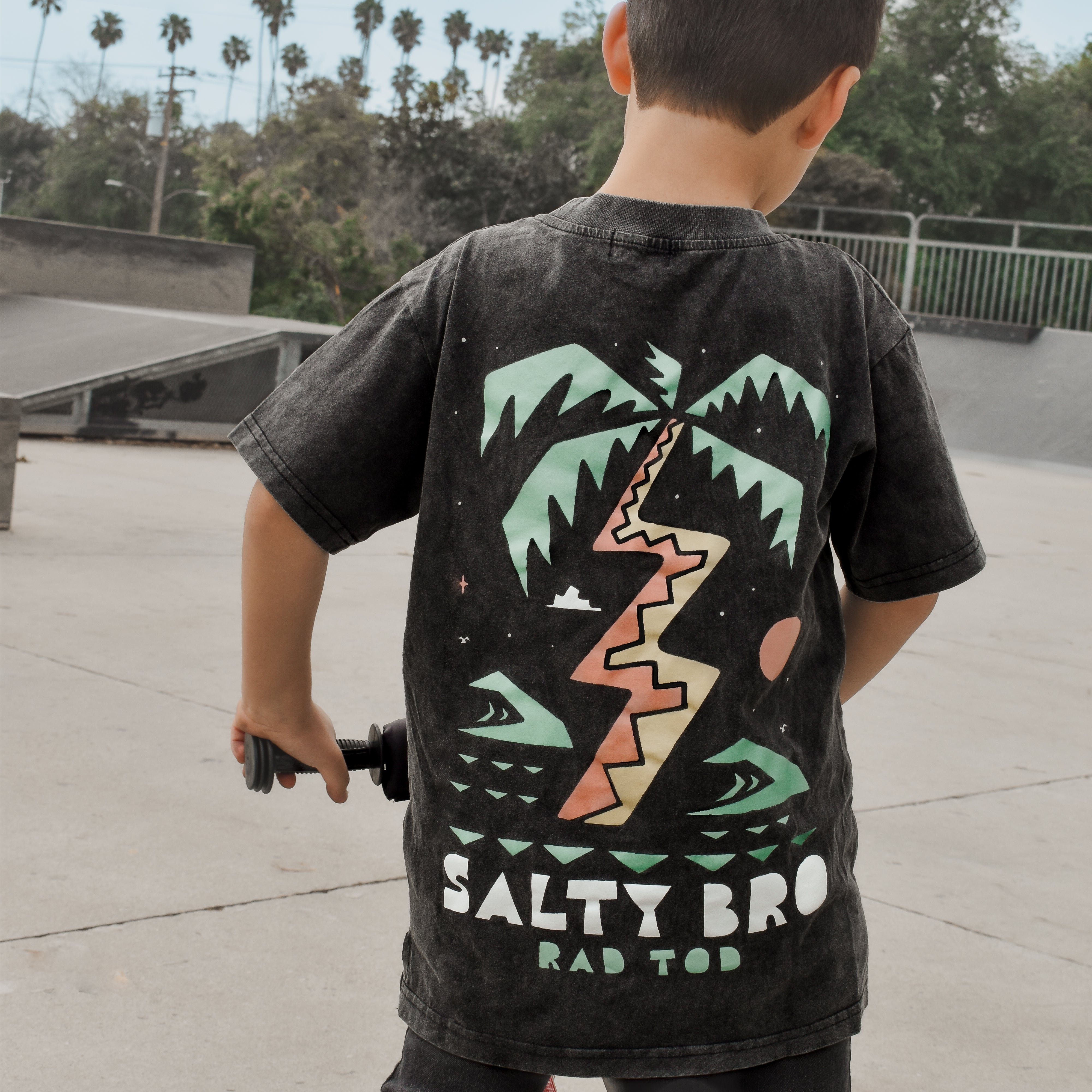 kid on bike wearing Rad Toddler black acid wash toddler tee with bold 'Salty Bro' text in playful, beach style lettering