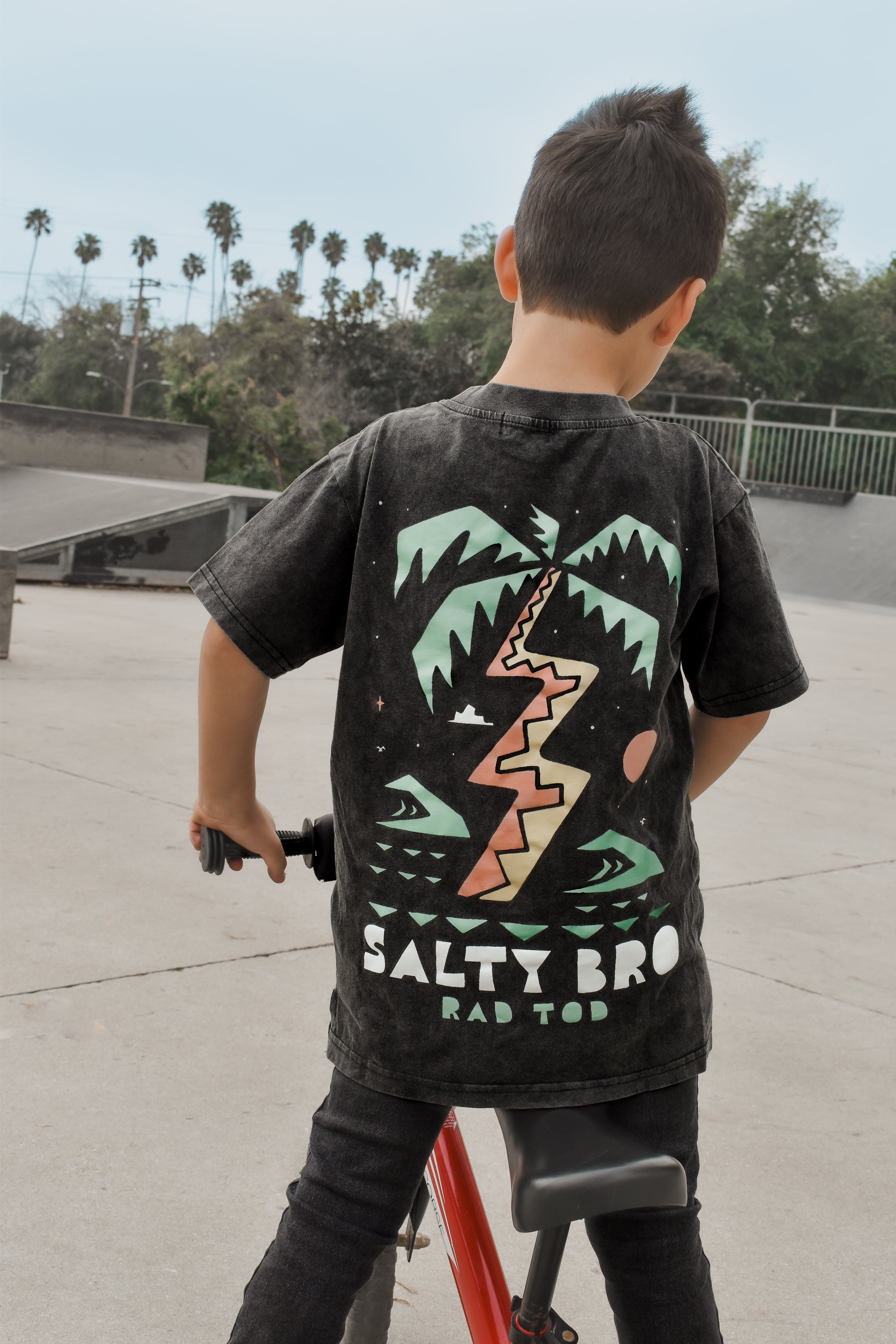 kid on bike wearing Rad Toddler black acid wash toddler tee with bold 'Salty Bro' text in playful, beach style lettering