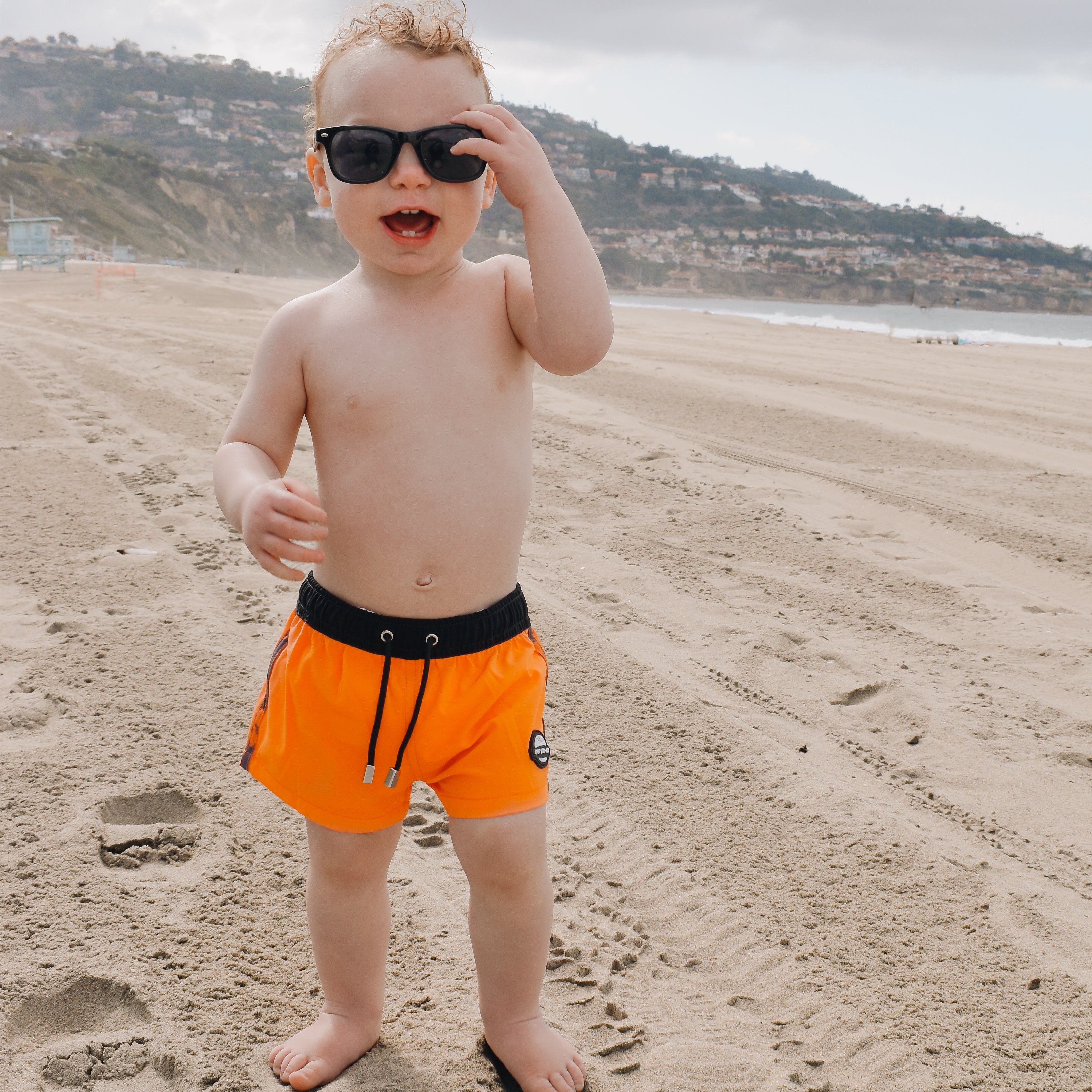 cute beach toddler wearing Bright swim shorts with an elastic waistband and drawstring, made from quick-drying fabric. Ideal for swimming, beach trips, and outdoor activities. Features vibrant patterns and a comfortable fit for active kids.