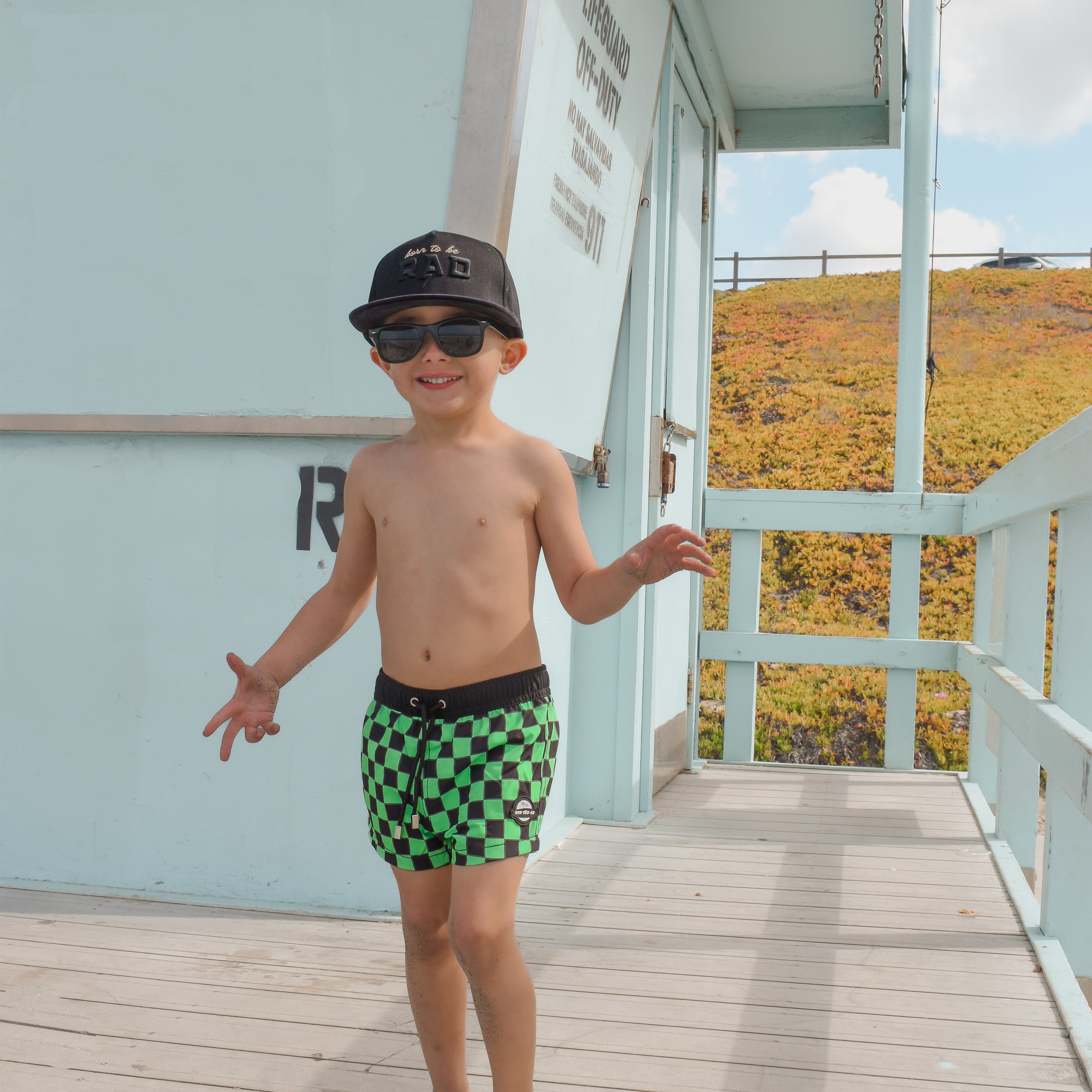 boy on lifeguard tower wearing Bright swim shorts with an elastic waistband and drawstring, made from quick-drying fabric. Ideal for swimming, beach trips, and outdoor activities. Features vibrant patterns and a comfortable fit for active kids.