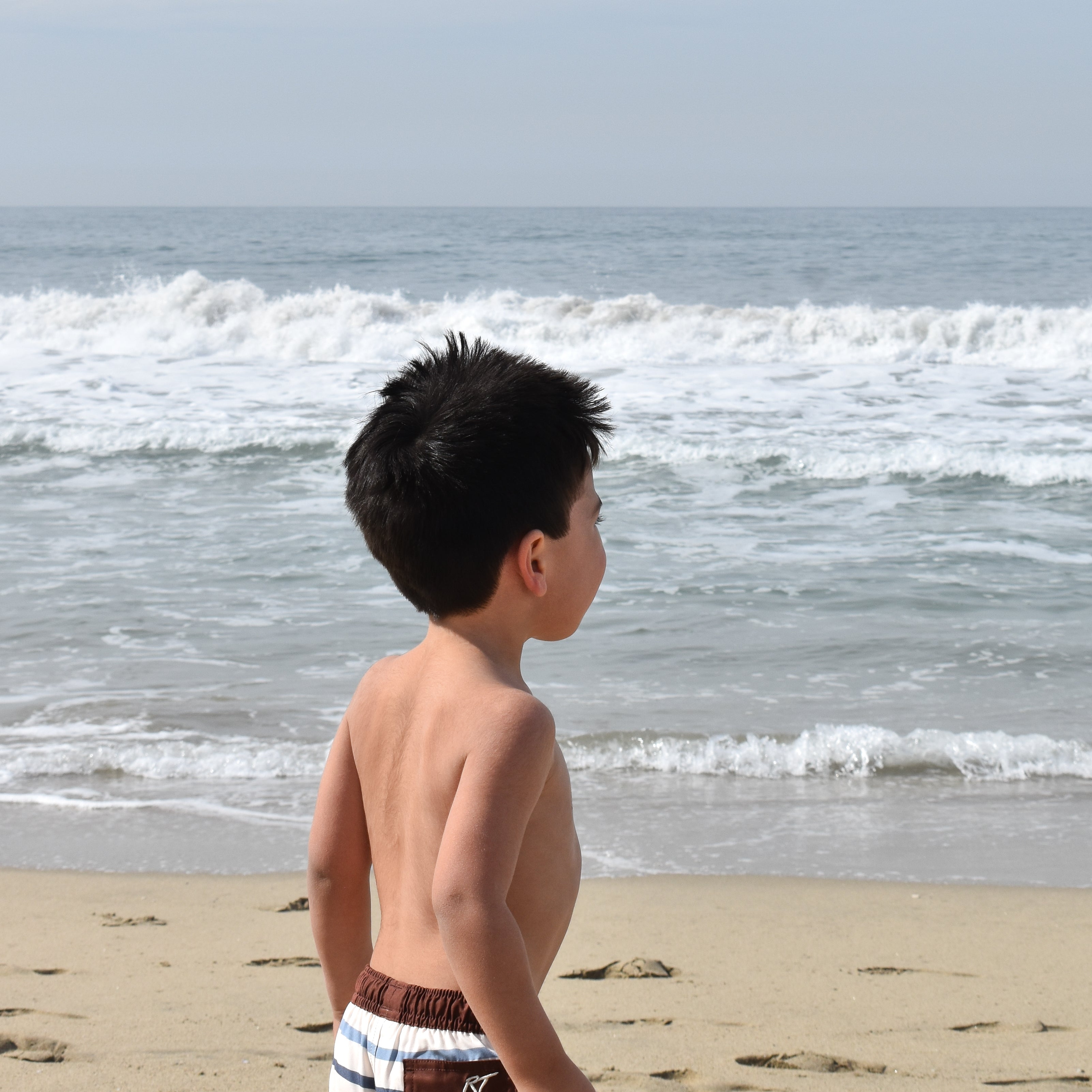 Young boy standing on a beach looking at the ocean wearing Driftwood Stripes by rad toddler