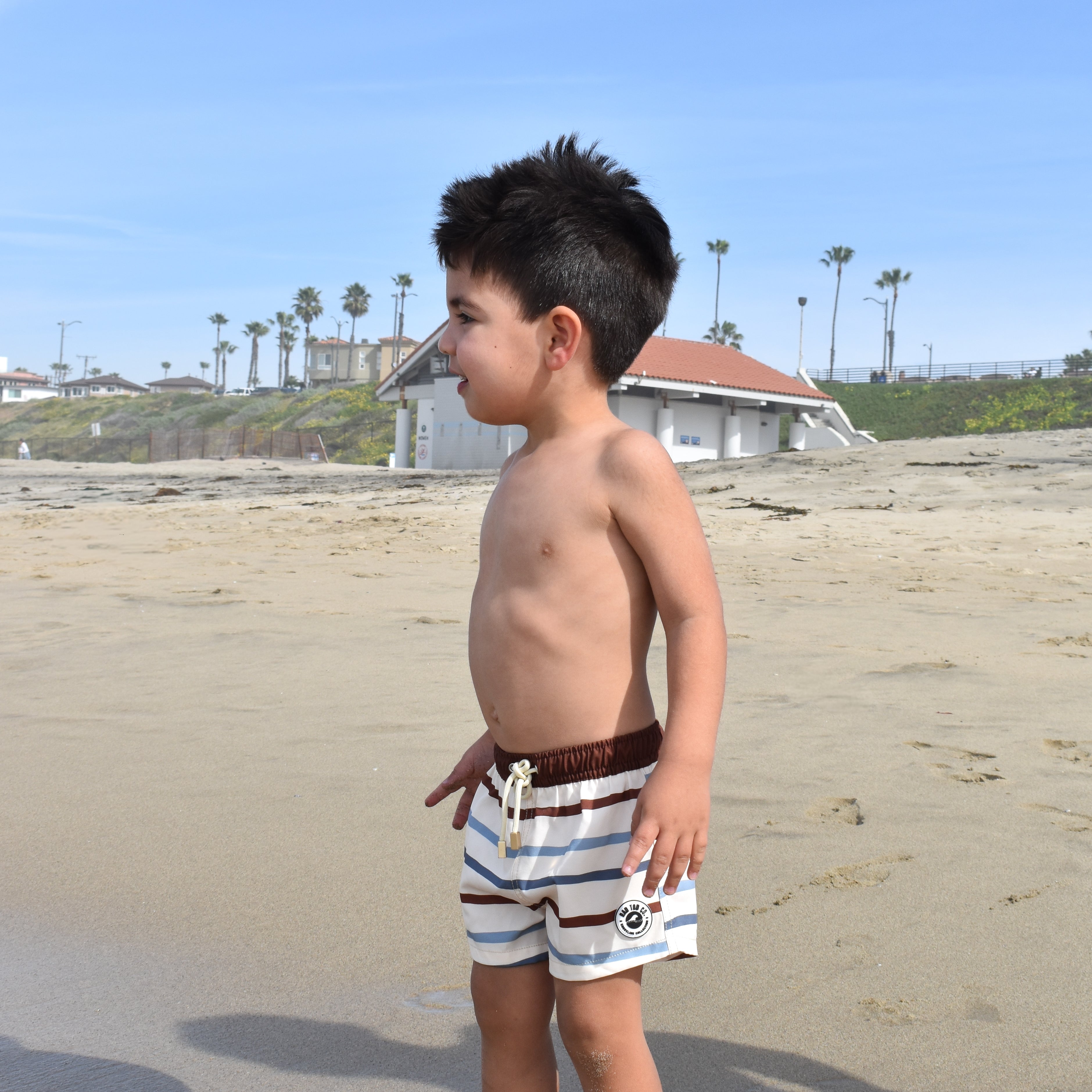 Young boy standing on a sandy beach with palm trees and a building in the background.
