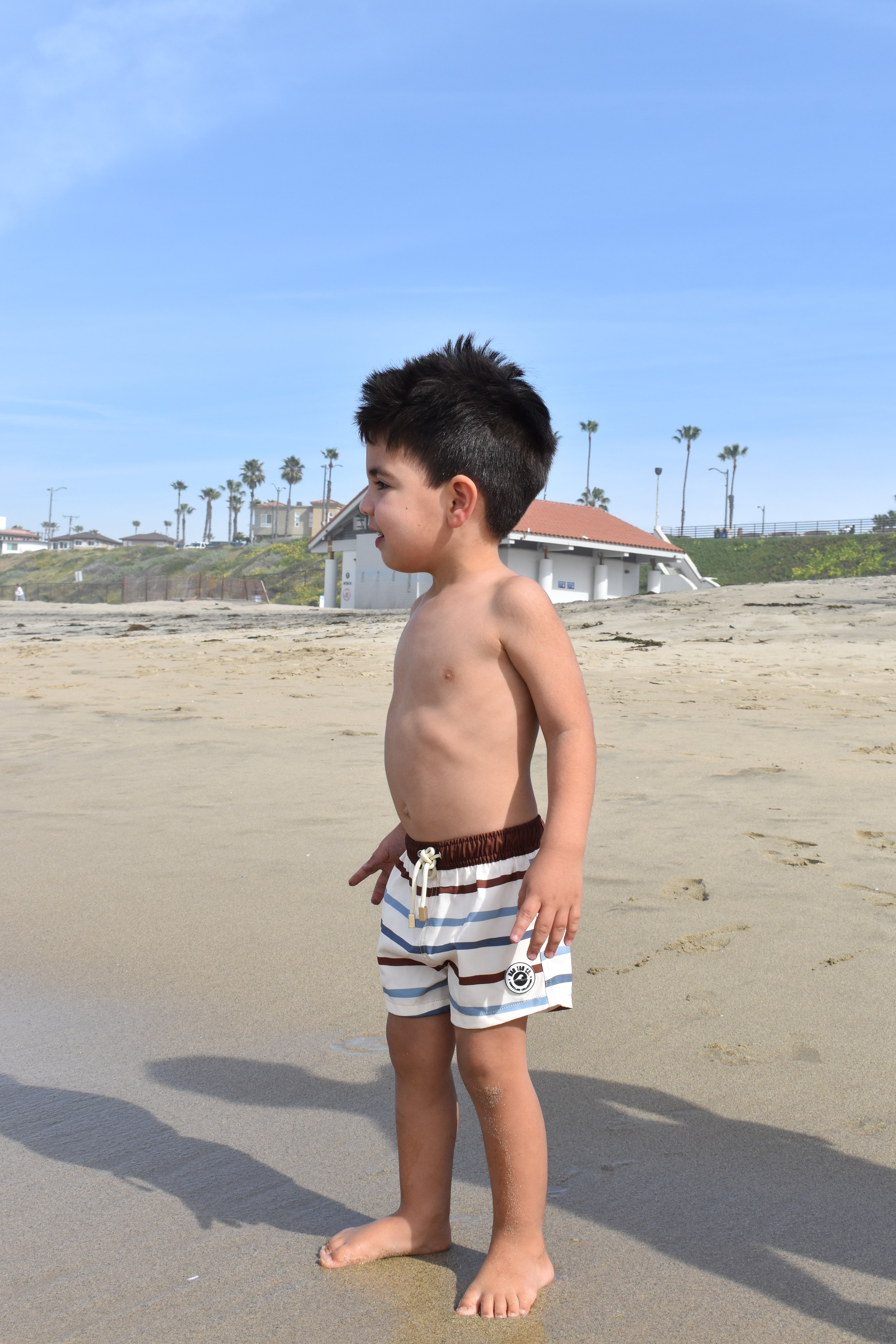 Young boy standing on a sandy beach with palm trees and a building in the background.