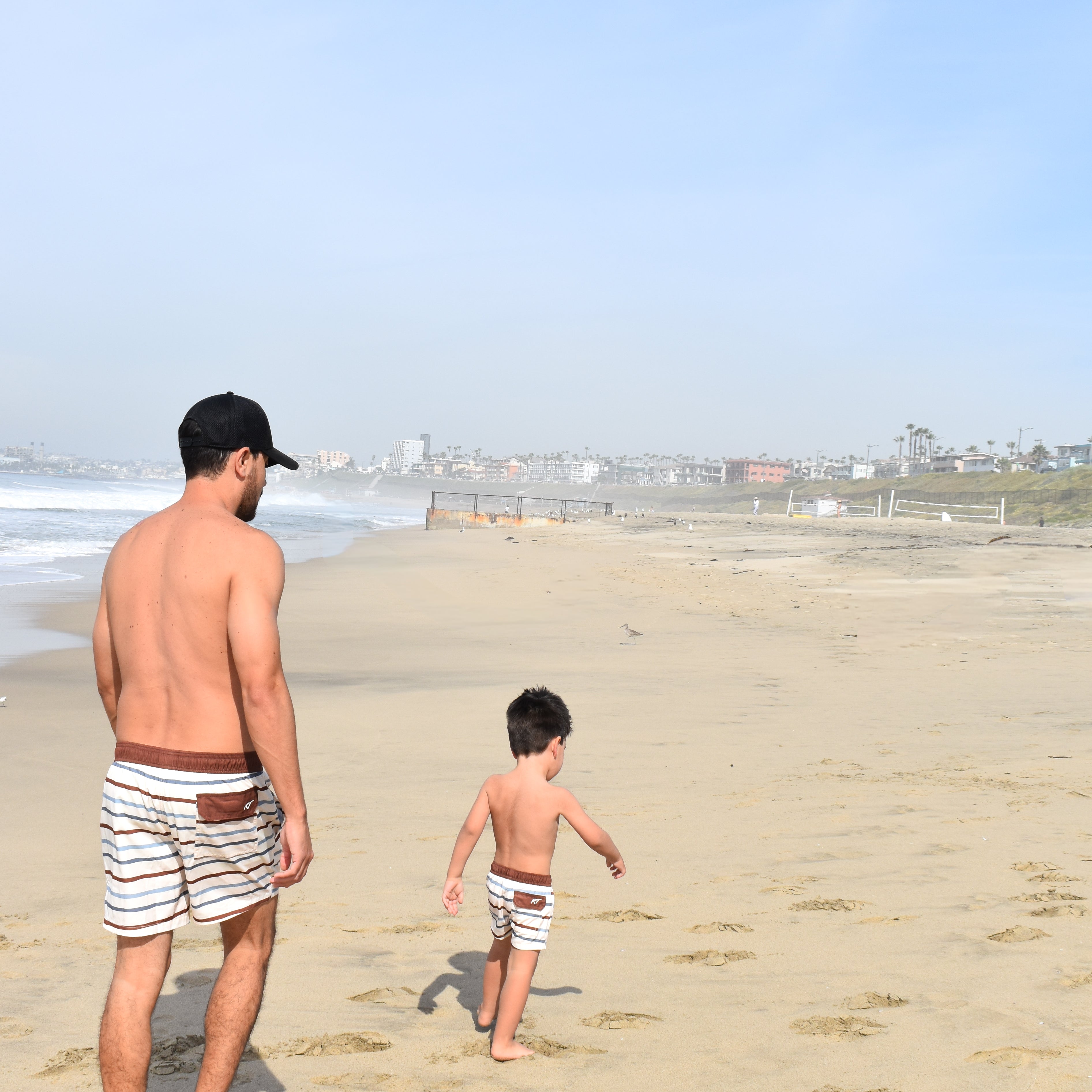 Man and child walking on a sandy beach with a clear sky wearing Driftwood Stripes matching swimmies by rad toddler