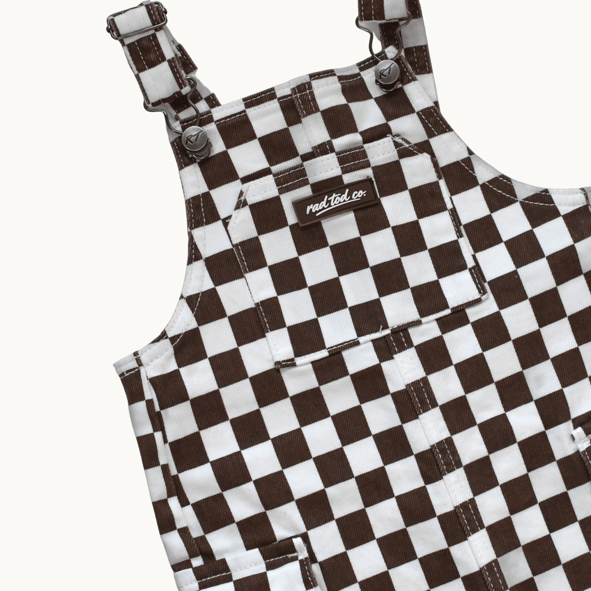 A close-up of a brown and white checkerboard patterned overall garment, showing the shoulder straps, front pockets, and a small visible Rad Tod brand label.