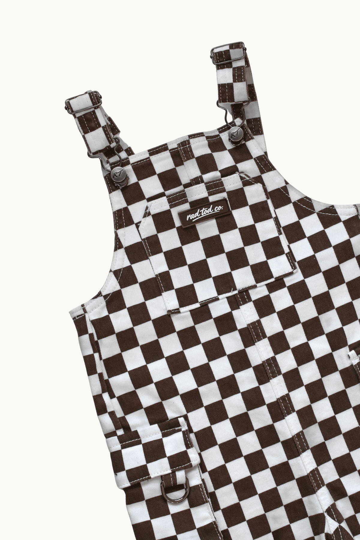 A close-up of a brown and white checkerboard patterned overall garment, showing the shoulder straps, front pockets, and a small visible Rad Tod brand label.