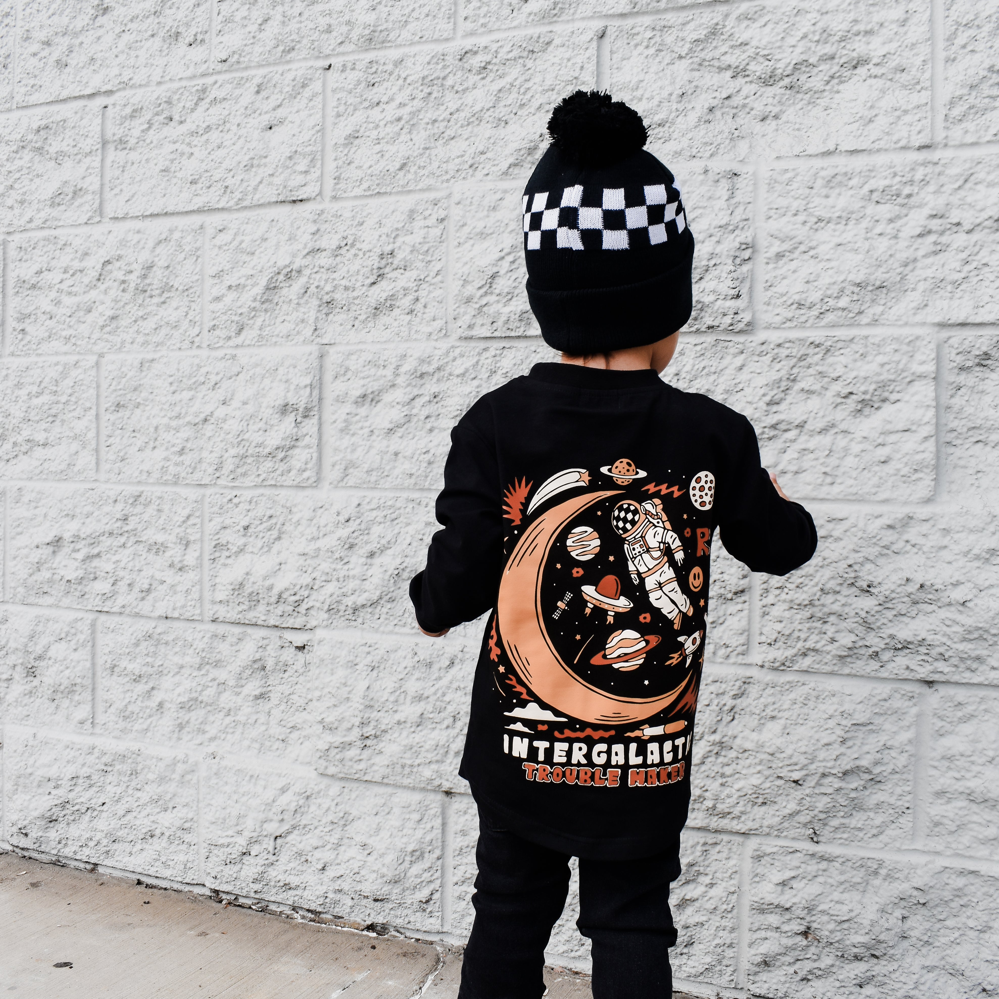Child wearing a black outfit with a celestial design, standing against a white brick wall.