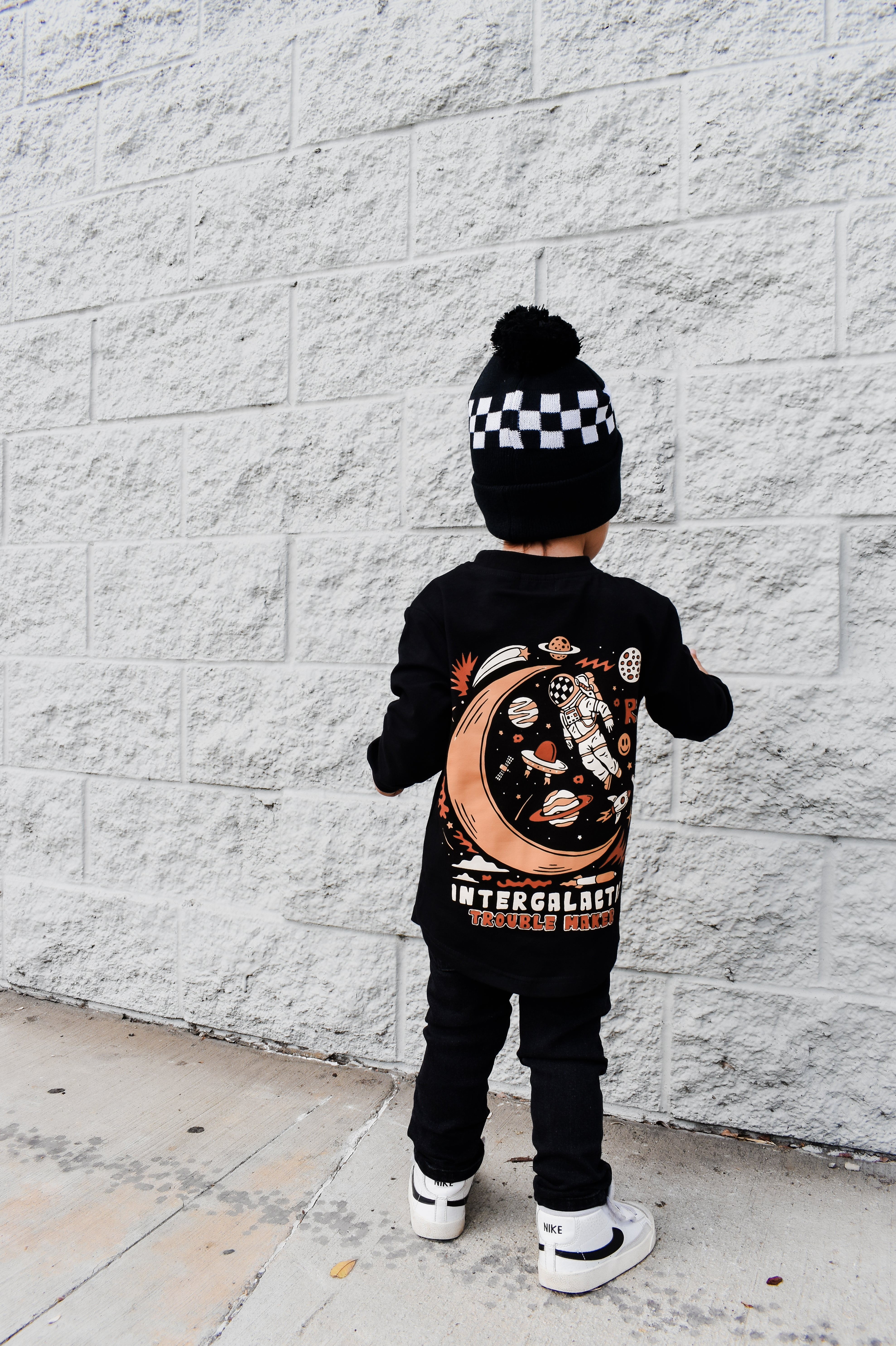 Child wearing a black outfit with a celestial design, standing against a white brick wall.