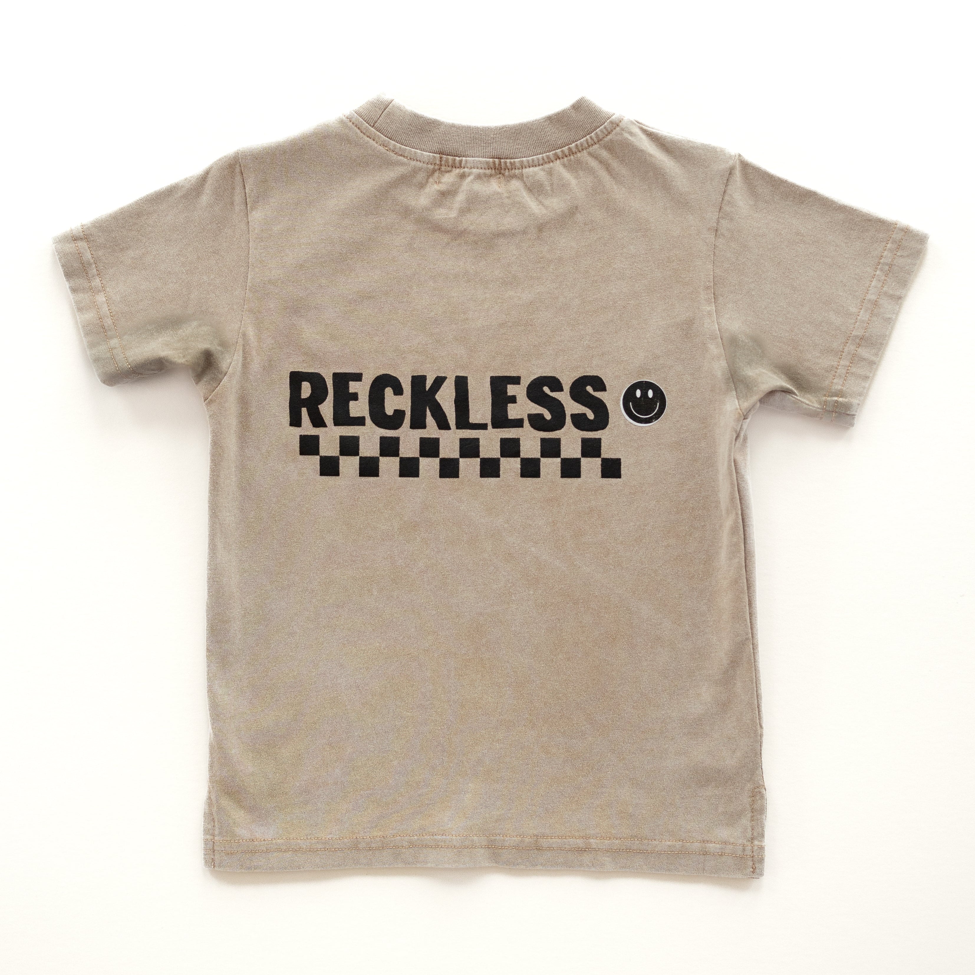 back side of Rad Toddler Beige acid wash toddler tee with bold 'Reckless' text, checkered patterns, and a smiley face graphic.