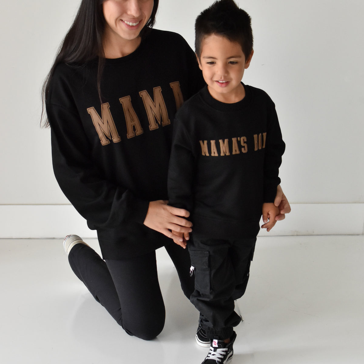 A woman and a young boy wearing matching black sweatshirts with gold 'MAMA' and 'MAMA'S BOY' text, posing together in a bright white studio setting.