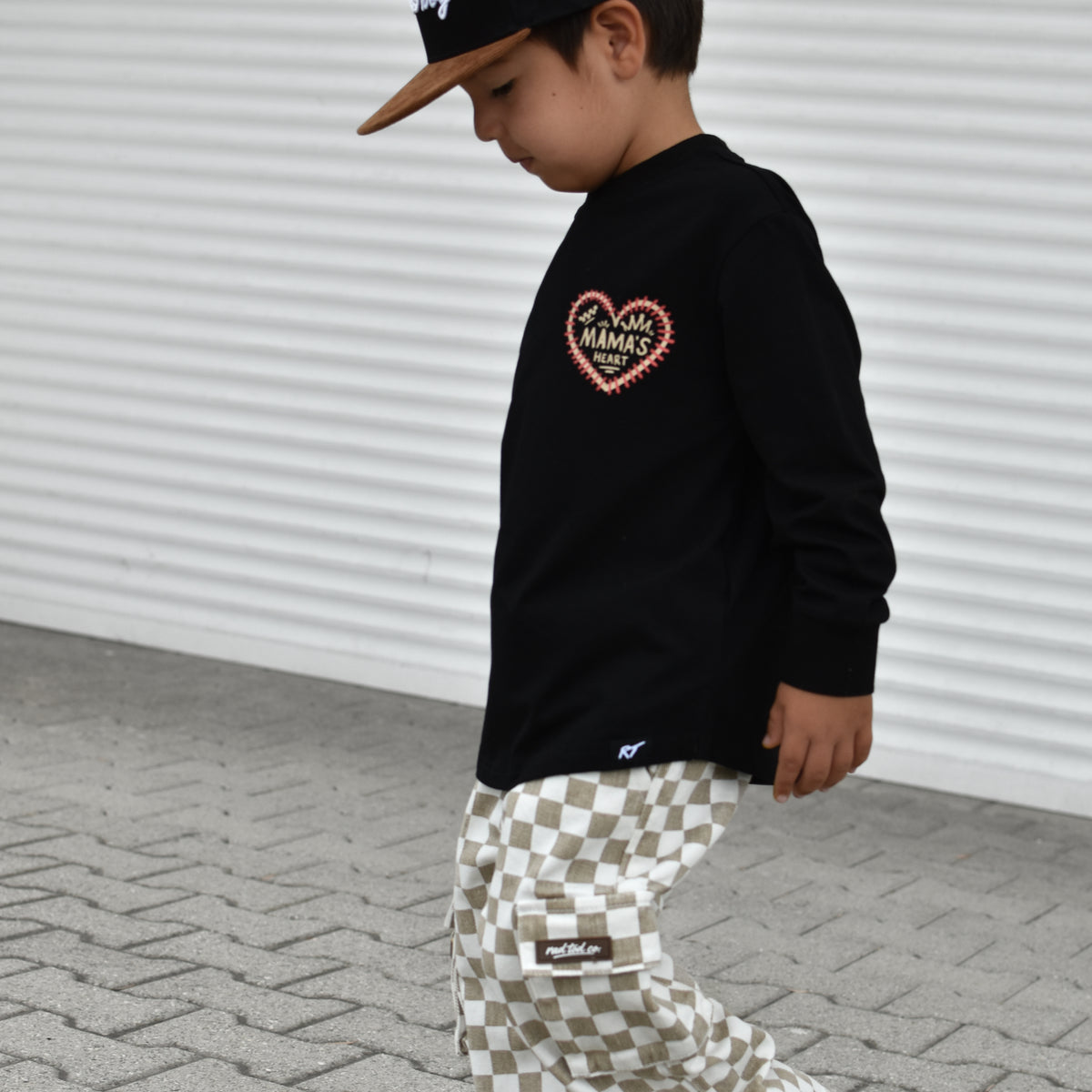 A child wearing a black long-sleeve shirt with a heart graphic, beige and white checkered pants, and a black cap, walking on a paved surface against a white corrugated wall.