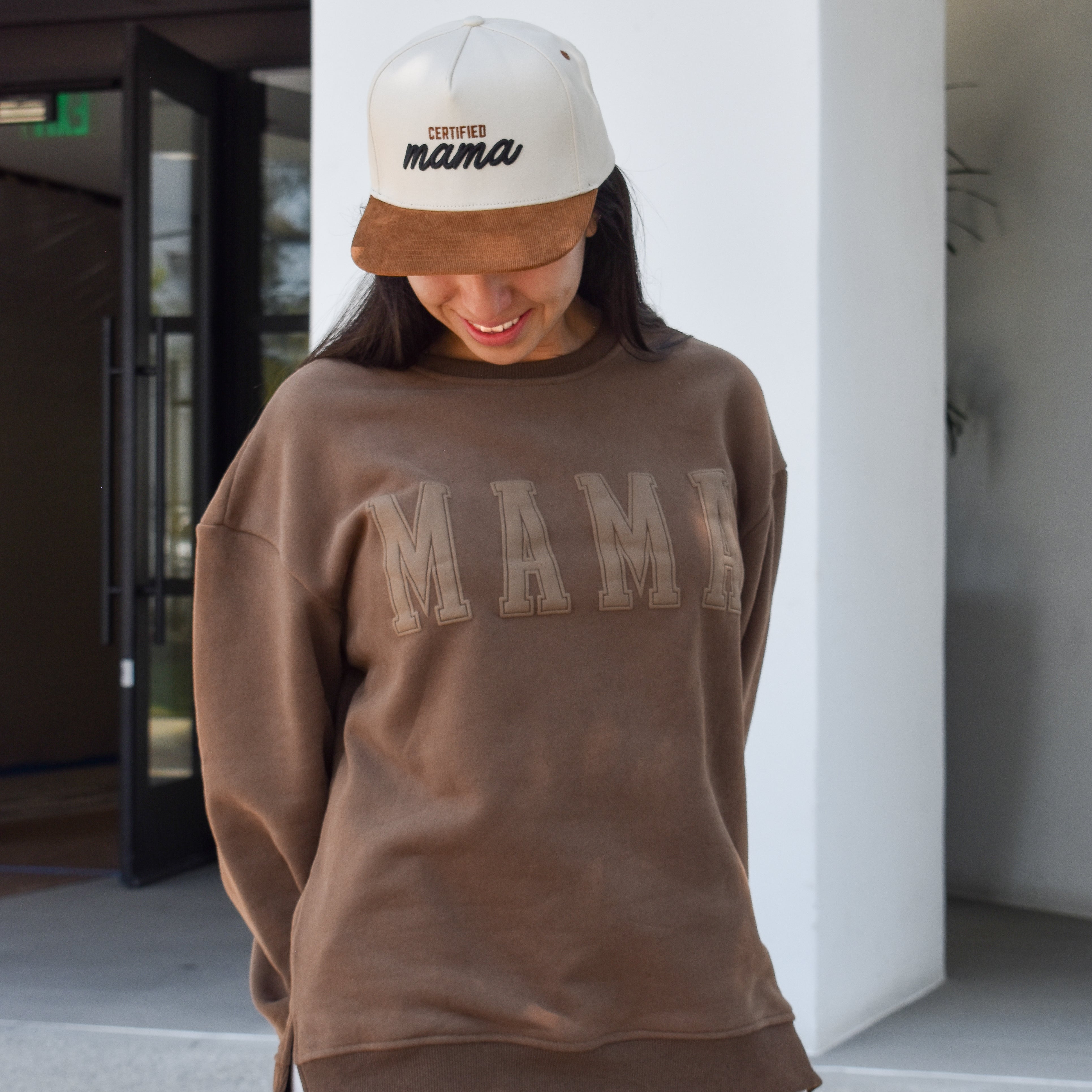 Person wearing a brown sweatshirt with 'MAMA' text, white pants, and a white cap