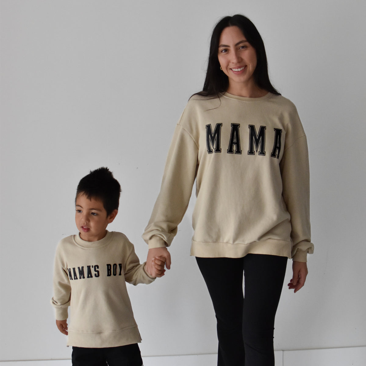 A woman and a young boy holding hands, both wearing matching beige crewneck sweatshirts with black lettering. The woman's sweatshirt reads 'MAMA' and the boy's reads 'MAMA'S BOY'.