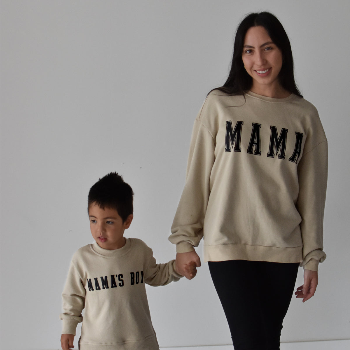 A woman and a young boy holding hands, wearing matching beige crewneck sweatshirts with black text. The woman's sweatshirt reads 'MAMA' and the boy's reads 'MAMAS BOY'.