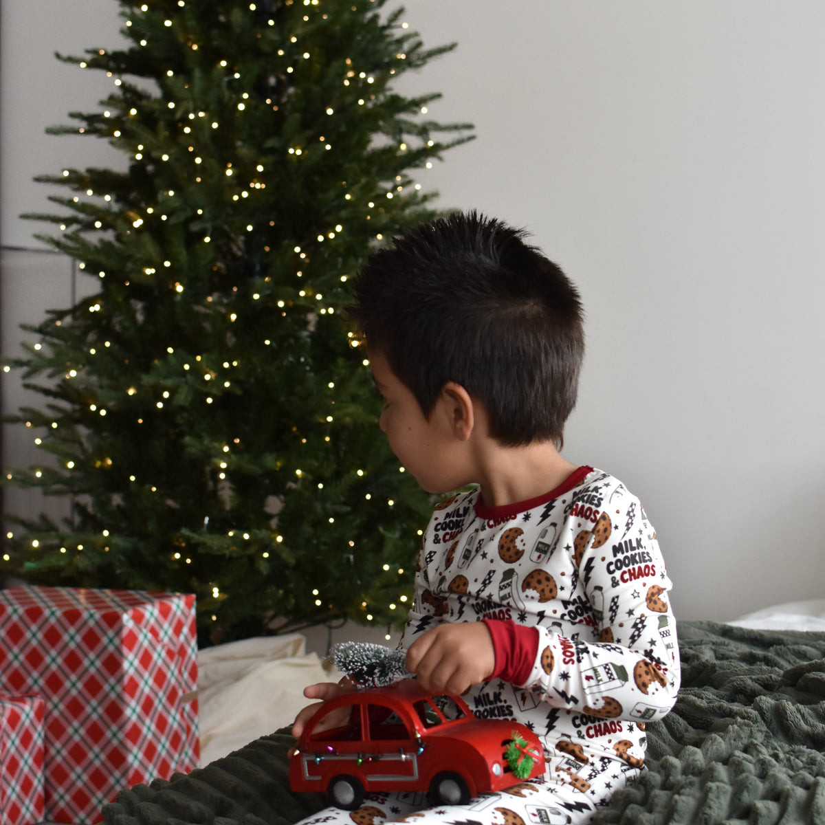 A young child wearing a white Christmas-themed pajama set with a holiday print sits on a dark green textured blanket in front of a lit Christmas tree, holding a red toy car.