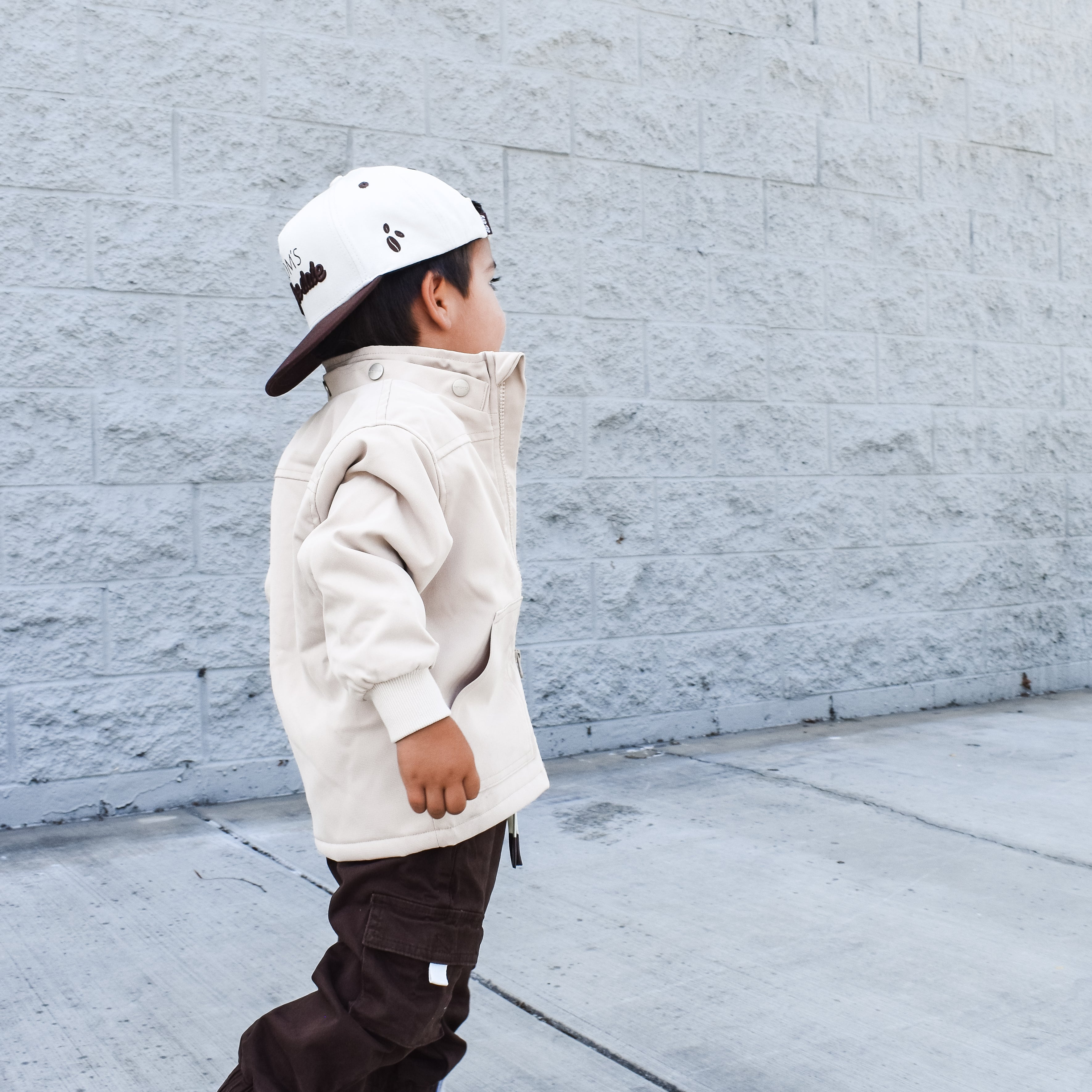 Child wearing a beige jacket, brown pants, and white sneakers against a gray wall.