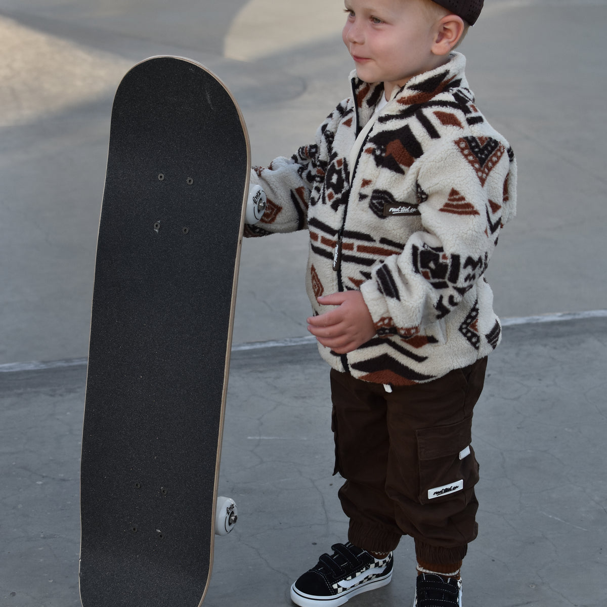 A young boy stands at a skatepark holding a skateboard, wearing a cream and brown patterned fleece jacket, dark brown pants, a black cap, and black sneakers.