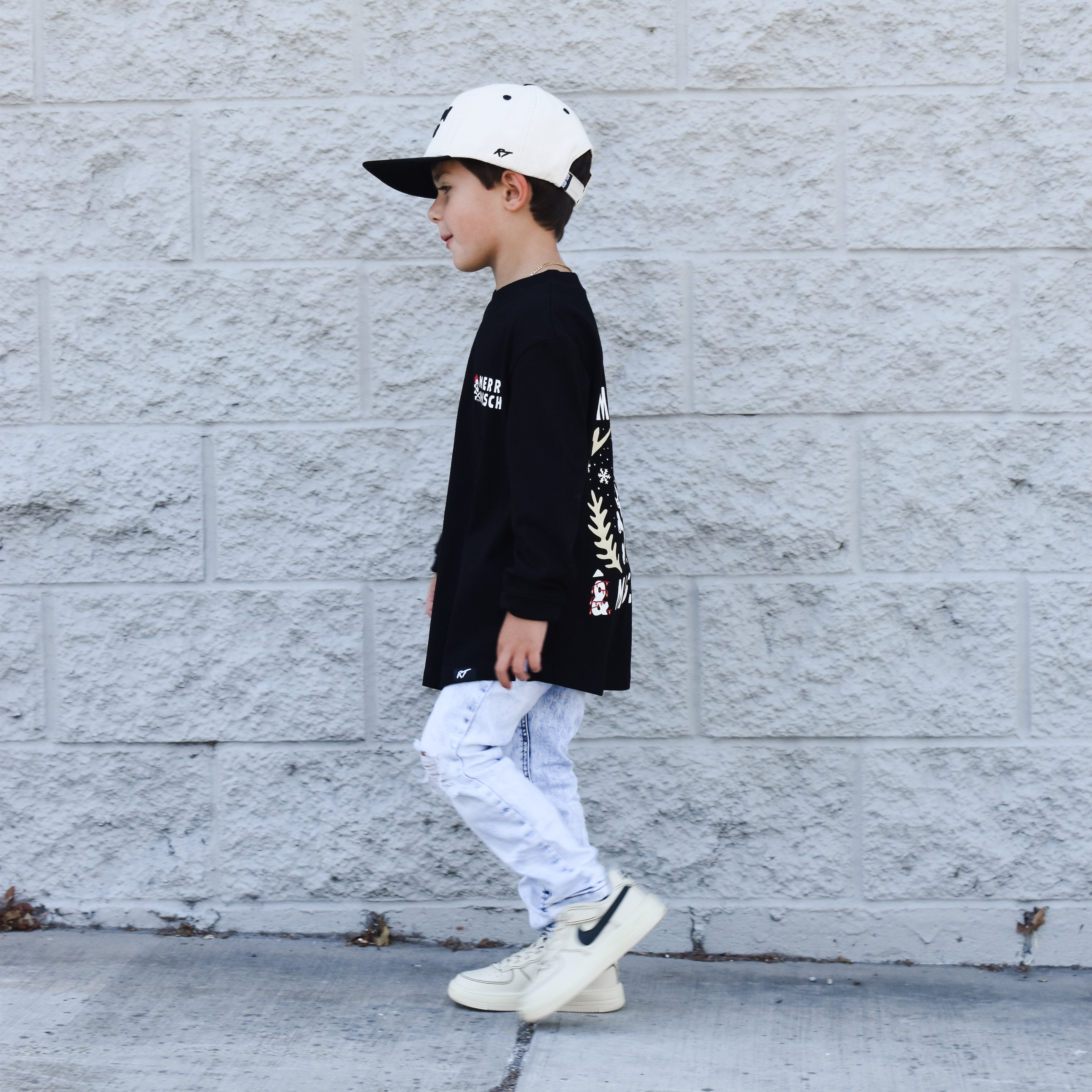 Child wearing a black long-sleeve shirt with white text, light blue jeans, and white sneakers against a gray wall.