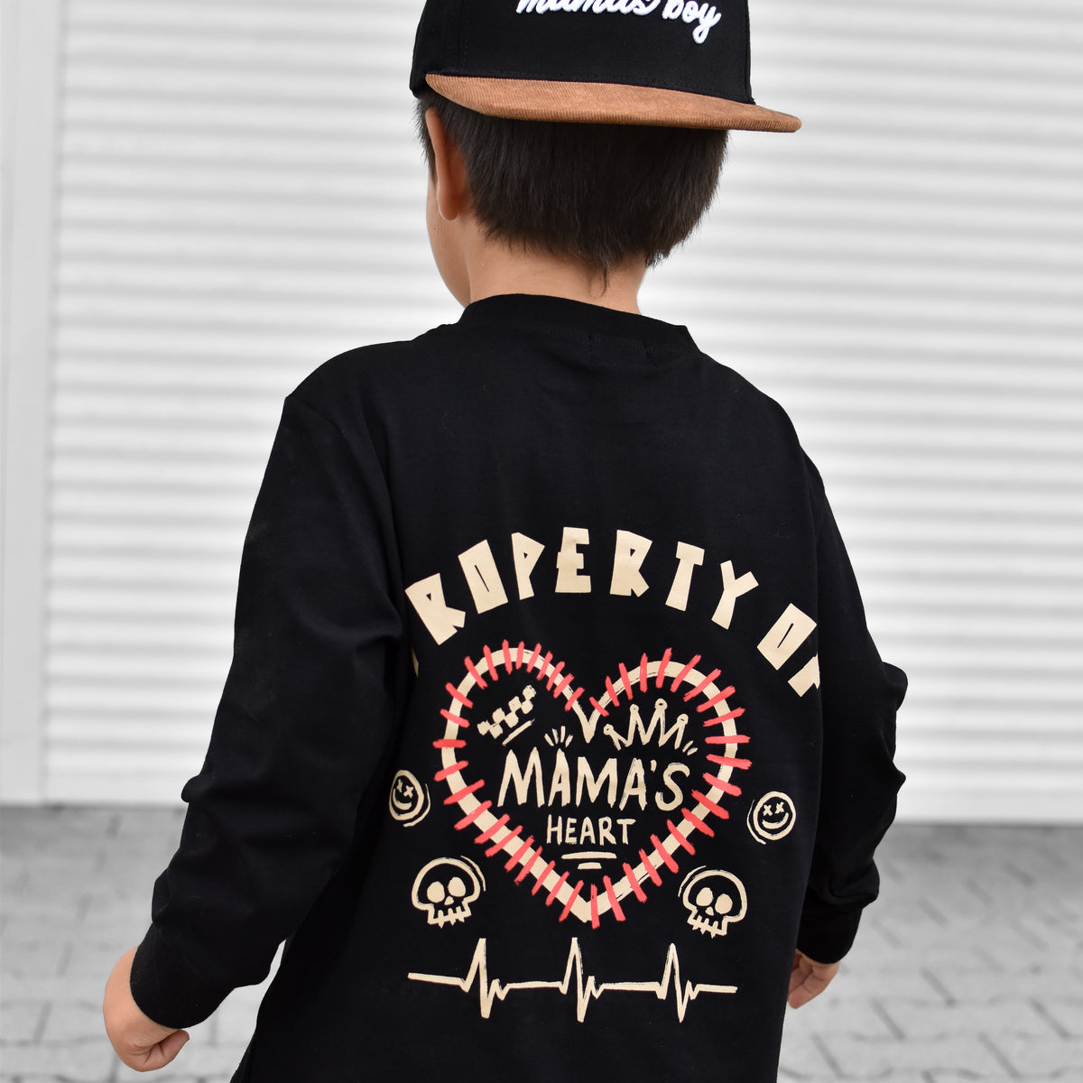 A child shown from behind wearing a black long-sleeve shirt with a large back graphic that reads 'PROPERTY OF MAMA'S HEART' in cream and red, along with a black cap that says ' Certified mamas boy'.