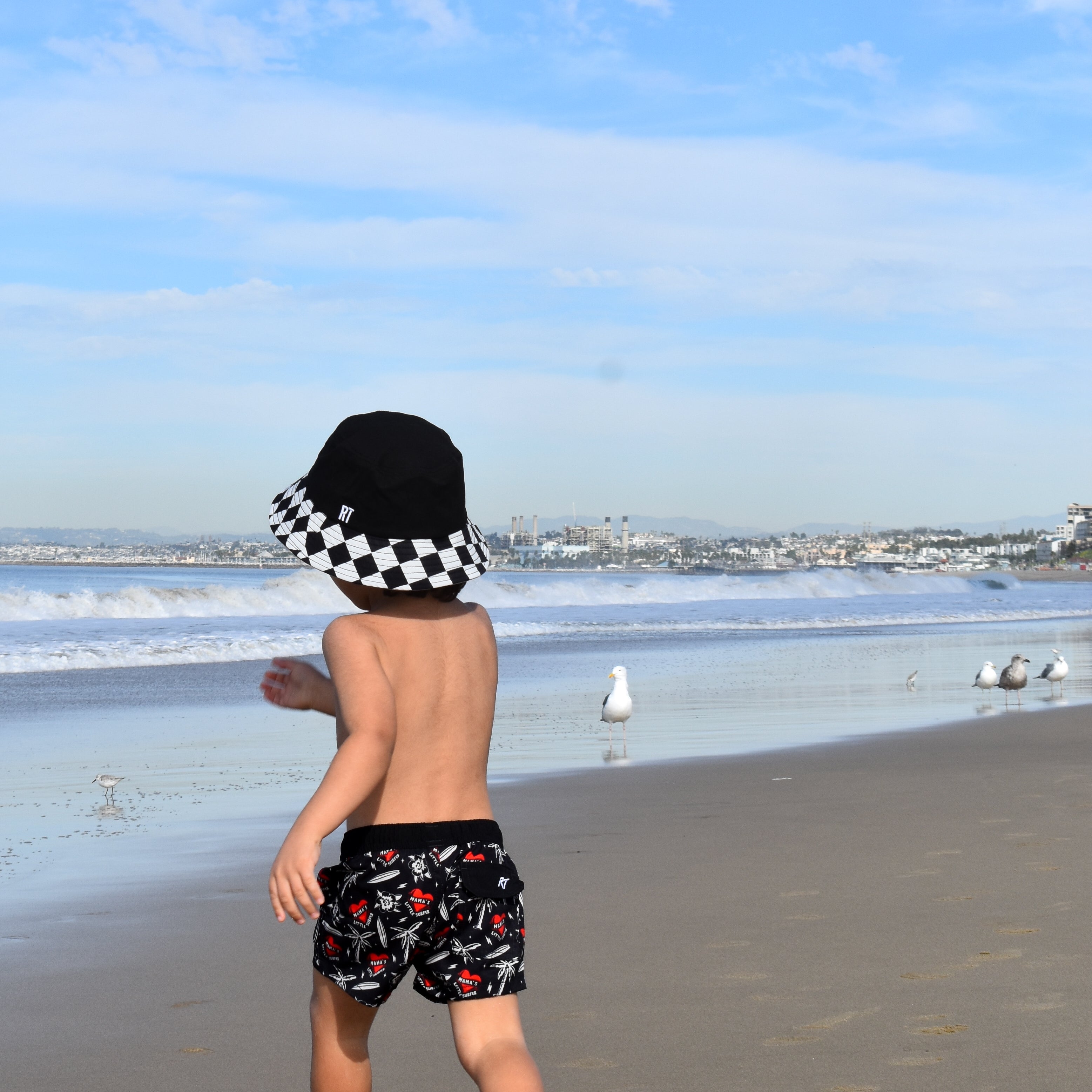 Child walking on a beach with a checkered hat and patterned shorts, facing the ocean.
