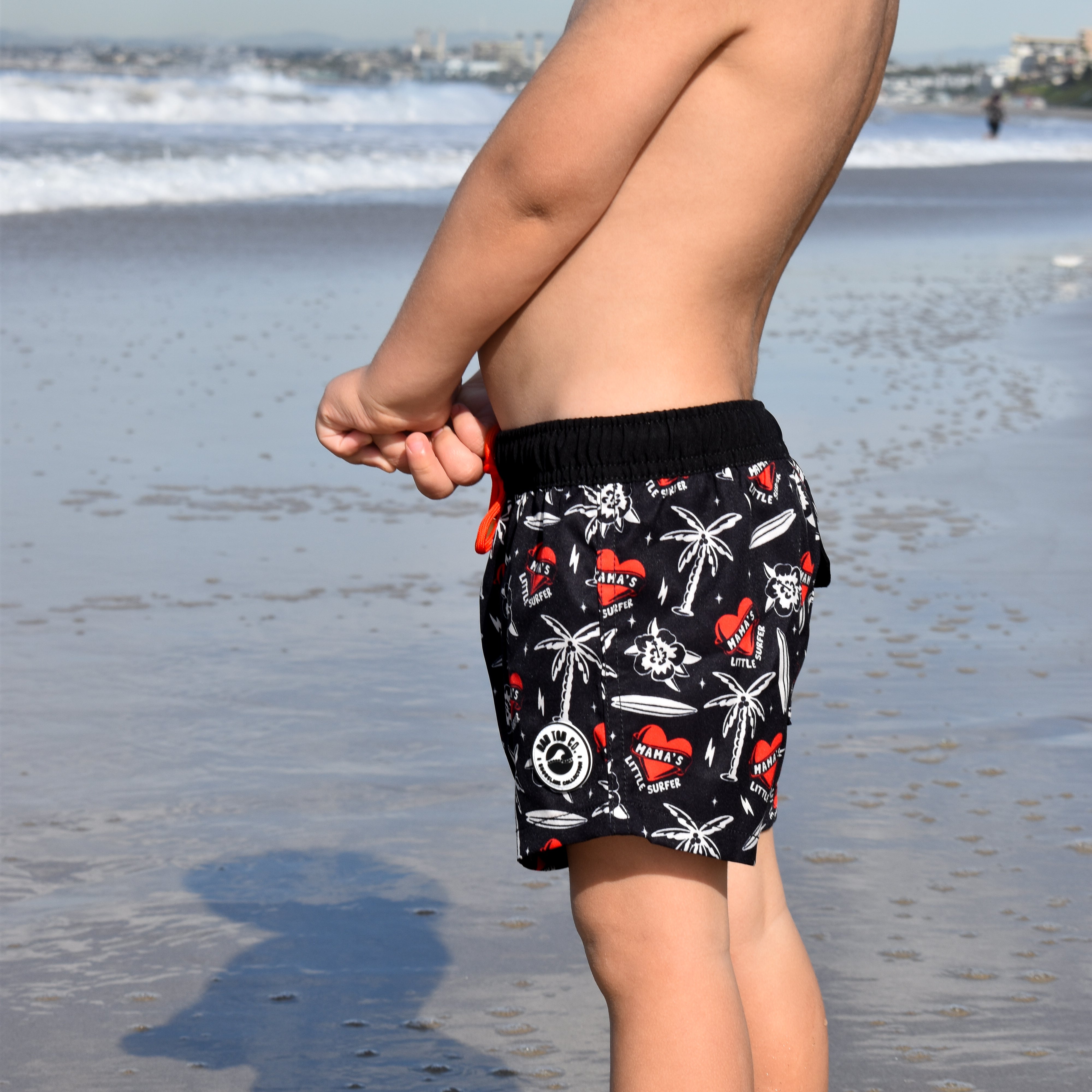 Toddler wearing Mama’s Little Surfer Swimmie in black with red heart and surf graphics at the beach