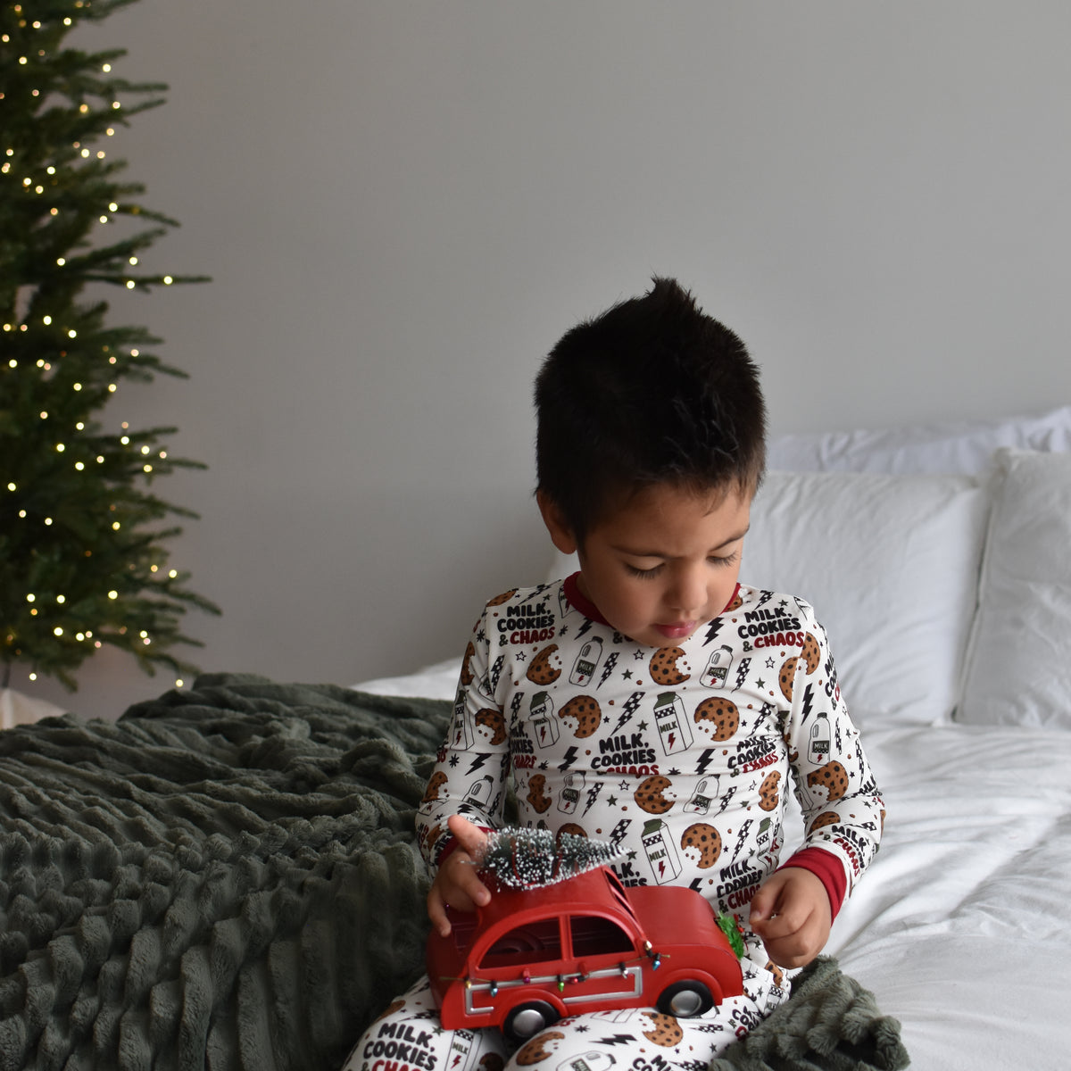 A young boy sits on a bed wearing white pajamas with a milk cookies and chaos print, holding a red toy car. A dark green textured blanket is draped over the bed, and a decorated Christmas tree is visible in the background against a gray wall.