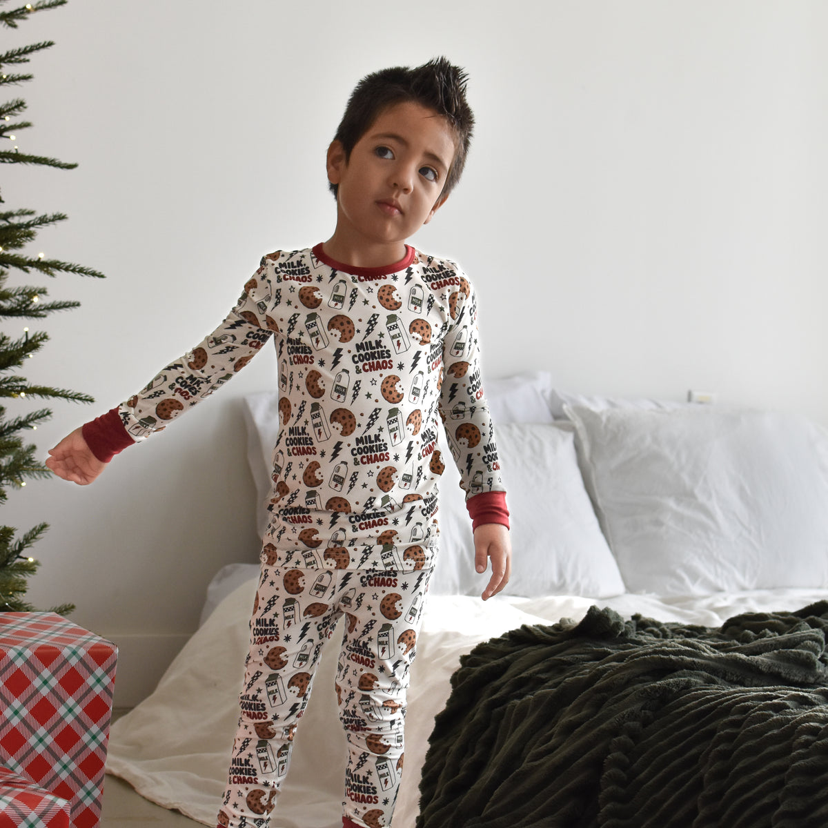 A young boy wearing a long-sleeve Christmas-themed pajama set with a brown and red milk cookies and chaos print by Rad TOddler on a white background, standing in a bedroom next to a bed and a Christmas tree.