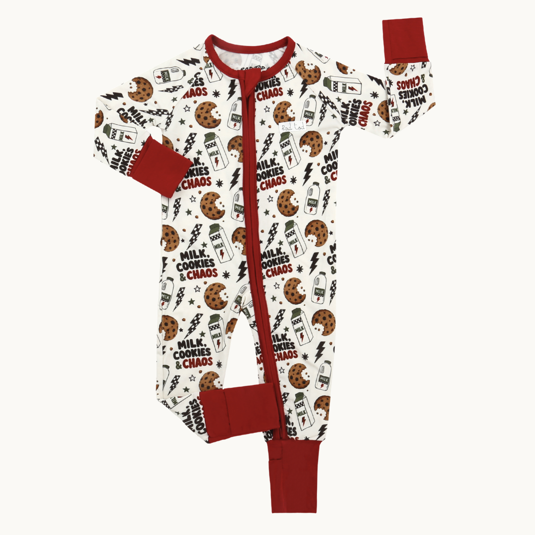 A baby's long-sleeve footless pajama one-piece with a red zipper, red collar, cuffs, and hem, featuring an all-over print of chocolate chip cookies, milk cartons, and the text 'MILK COOKIES & CHAOS' on a cream background.